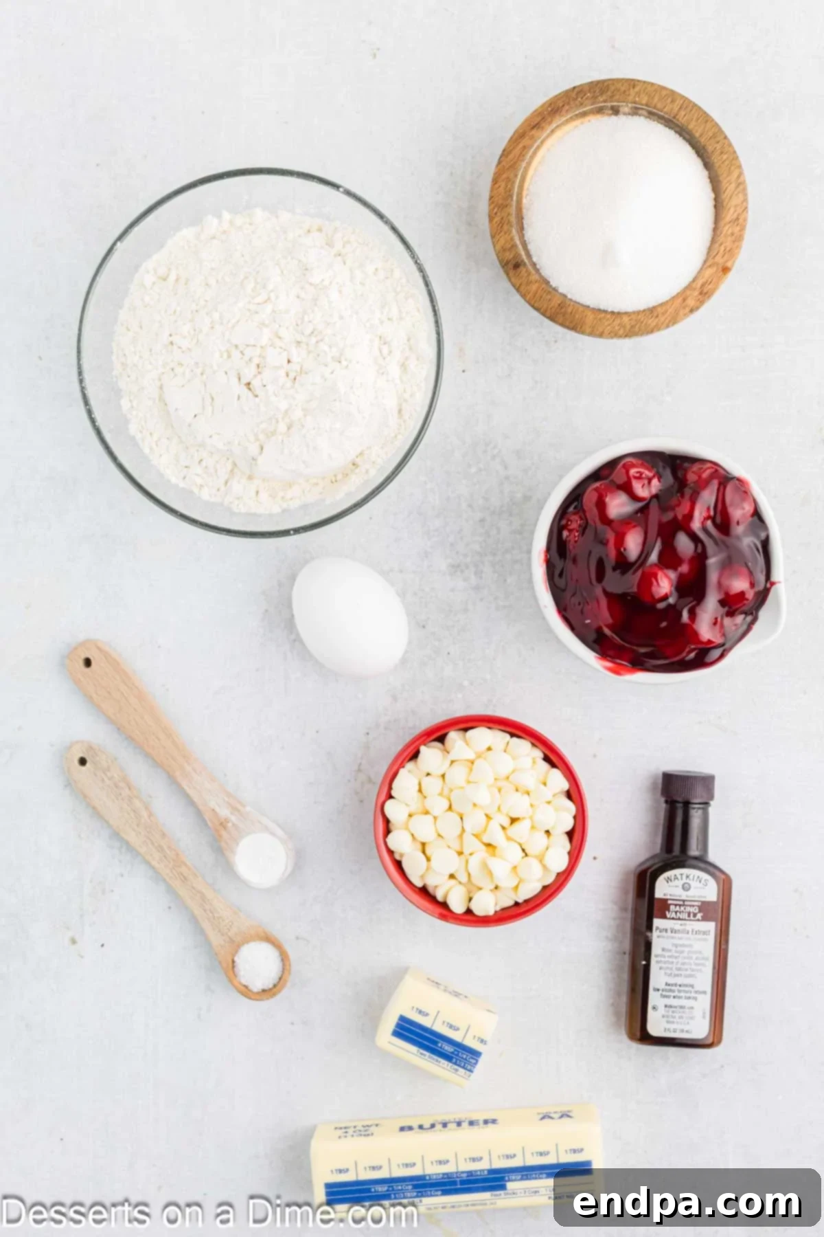 Crimson Cherry Thumbprint Cookies 3 A flat lay photograph showcasing the ingredients needed for Cherry Thumbprint Cookies, including flour, baking powder, salt, softened butter, granulated sugar, an egg, vanilla extract, cherry pie filling, and white chocolate chips.