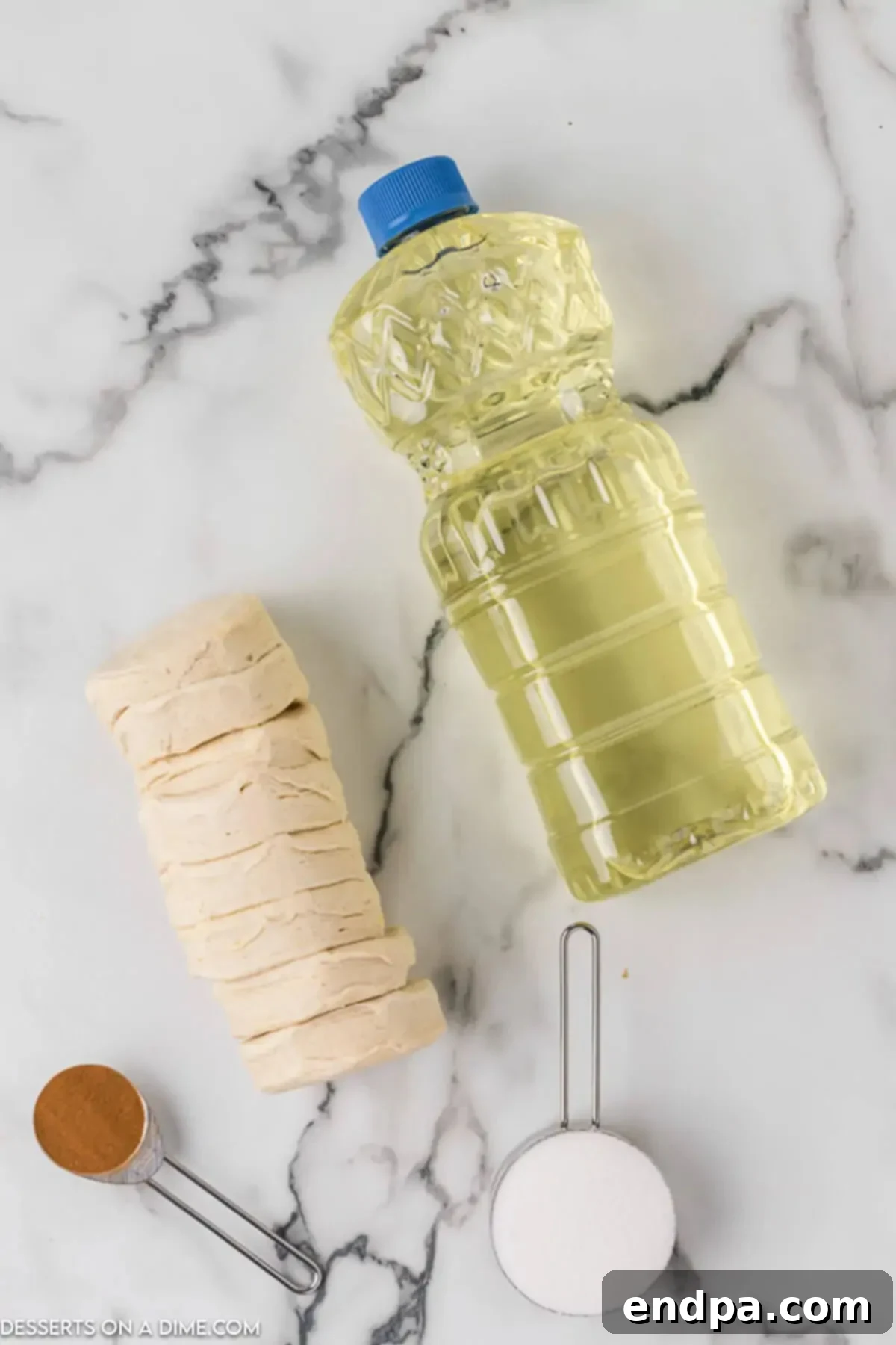 Four primary ingredients for canned biscuit donuts laid out on a kitchen counter: a tube of refrigerated biscuits, a bottle of vegetable oil, a bowl of white sugar, and a small container of ground cinnamon.
