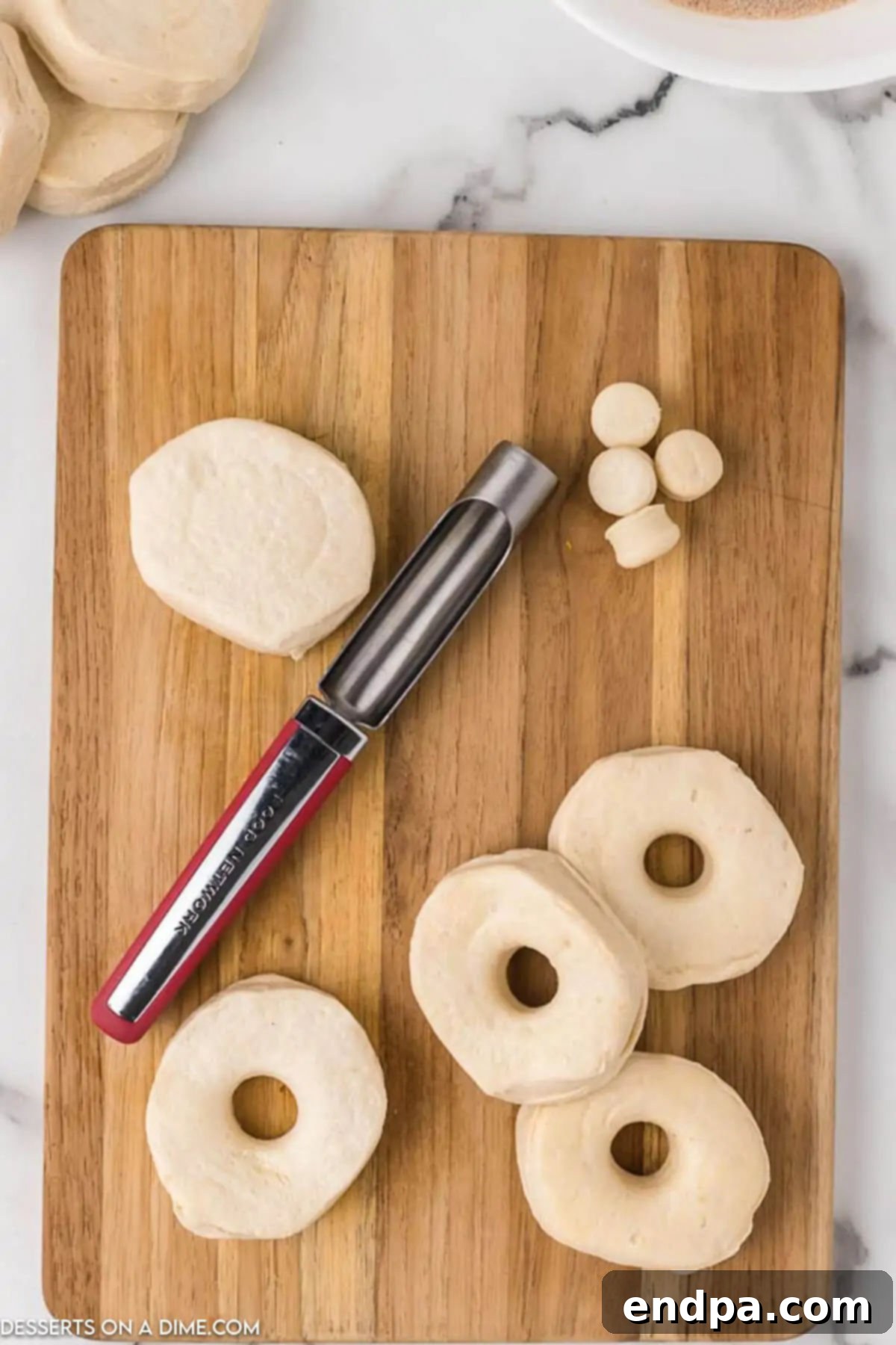 A hand using an apple corer to expertly cut a circular hole in the center of a raw refrigerated biscuit dough, creating the classic donut shape.