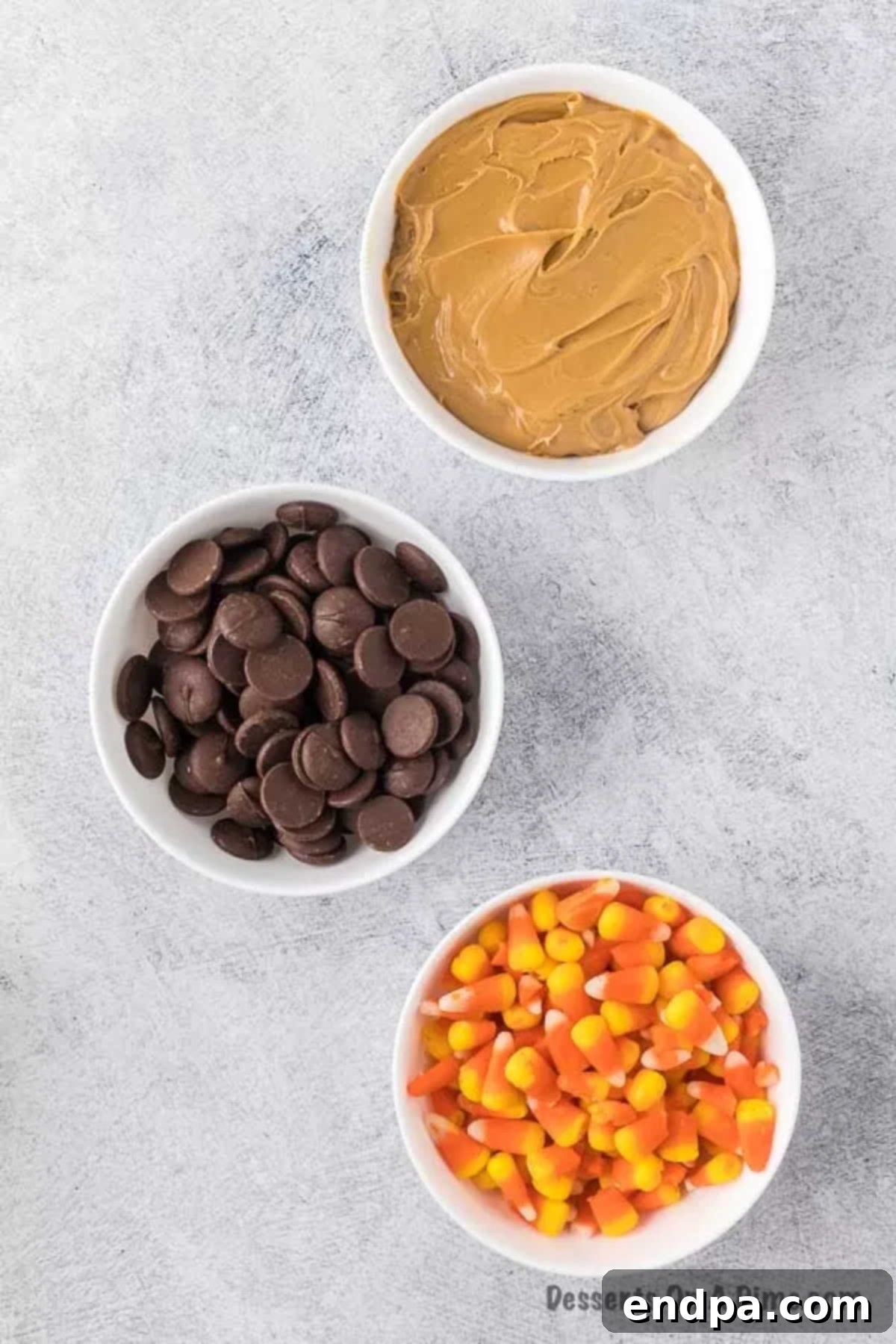 DIY Butterfinger Bliss 3 Close up shot of the three simple ingredients for homemade Butterfingers: a bowl of candy corn, a jar of creamy peanut butter, and a bag of chocolate candy melts.