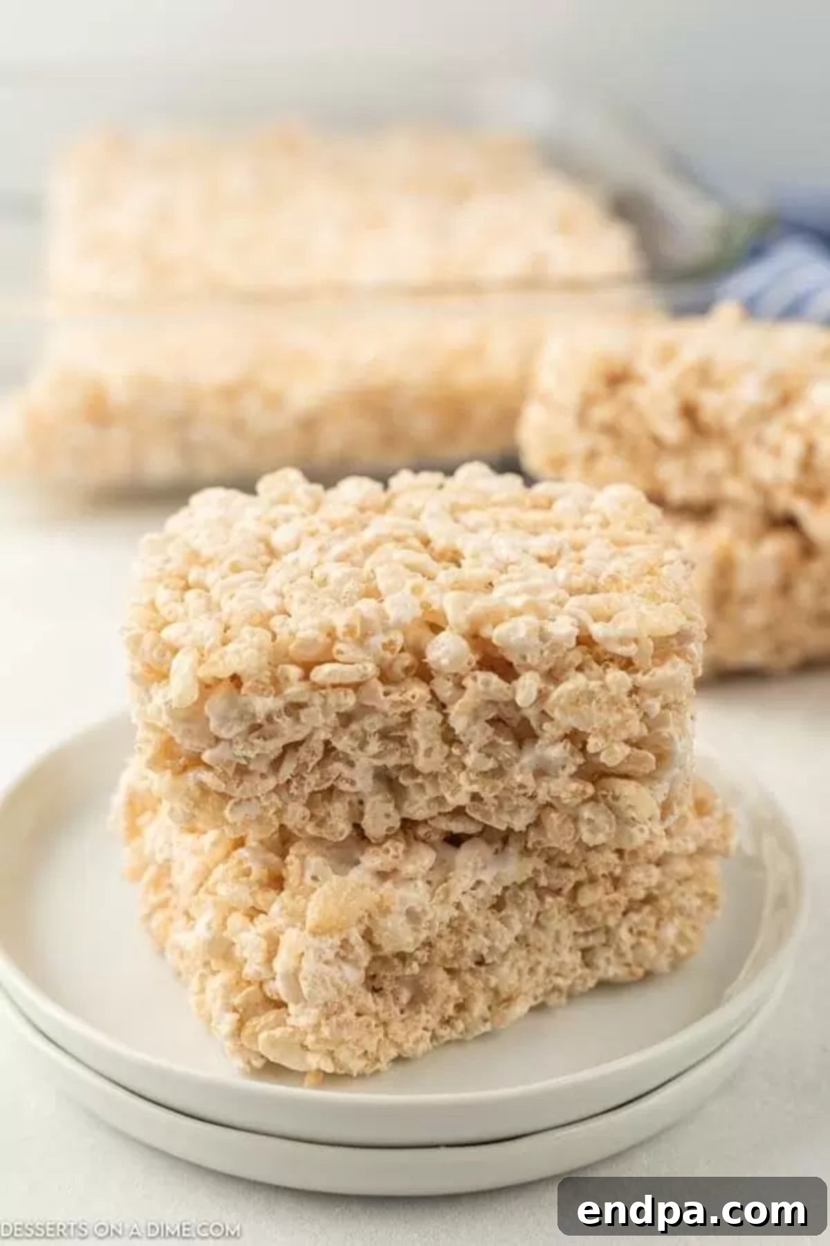 Cloud-Like Marshmallow Fluff Krispie Treats 2 Perfectly cut Rice Krispie treat squares arranged neatly on a white plate, showcasing their gooey texture.