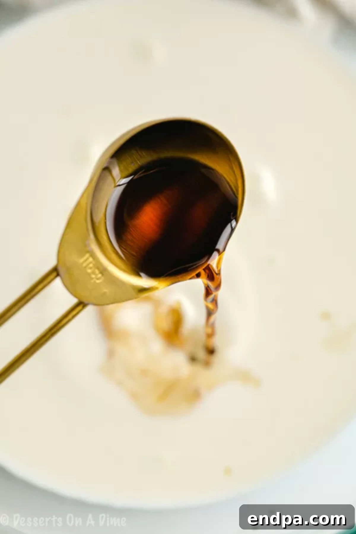 A measuring spoon pouring vanilla extract into a mixing bowl with heavy cream and erythritol.