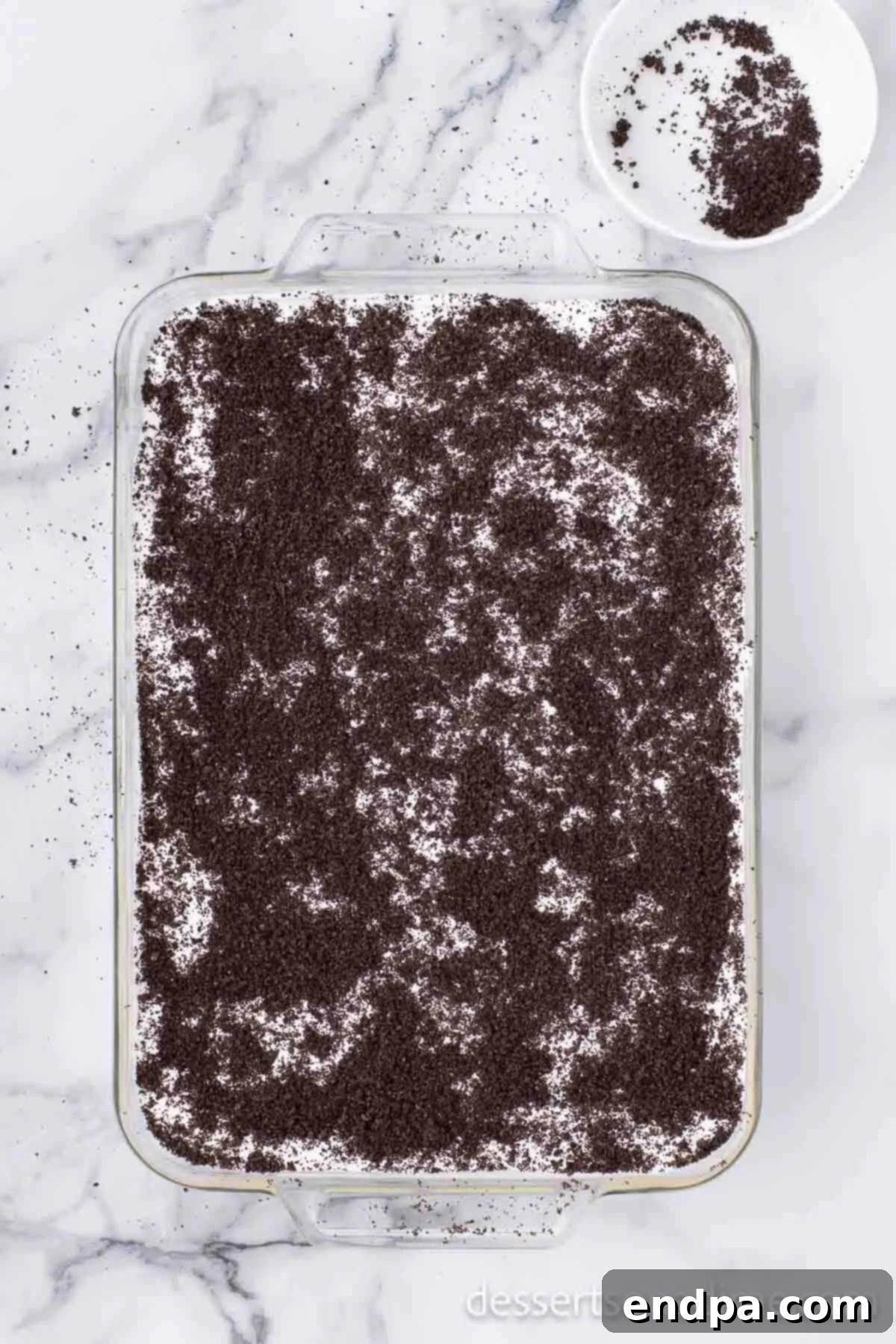 The top layer of crushed Oreo cookies being sprinkled over the whipped topping, completing the Dirt Cake assembly.