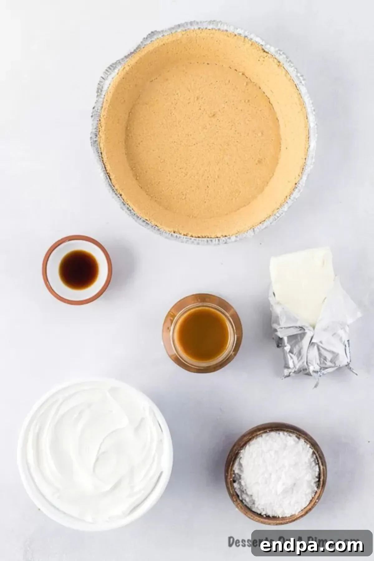 A spread of ingredients including a graham cracker crust, cream cheese, salted caramel sauce, and whipped topping, ready for making the no-bake pie.