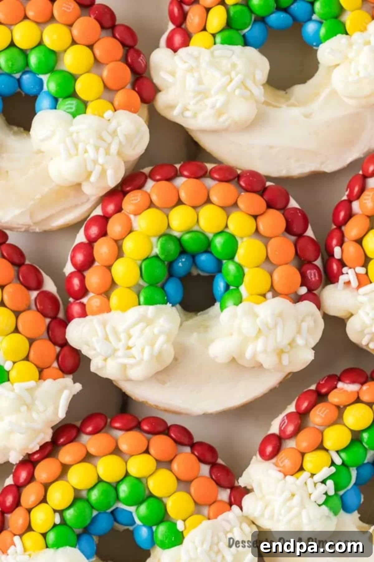 A vibrant display of homemade Rainbow Donuts, each decorated with a colorful candy rainbow and fluffy white frosting clouds, perfectly lined up.