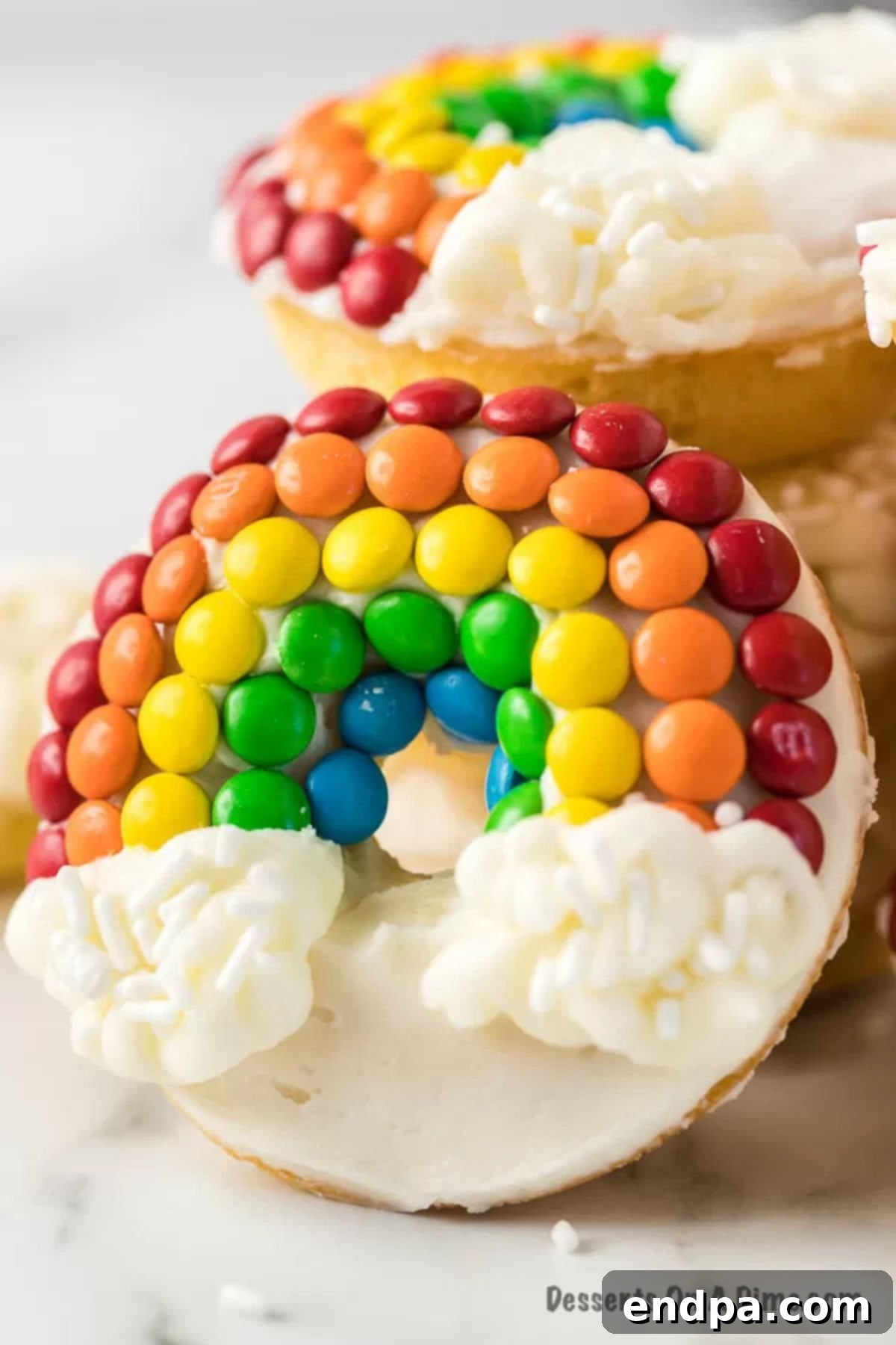 Another close-up of a Rainbow Donut, leaning against a small pile of donuts, showcasing its vibrant colors and cloud frosting.