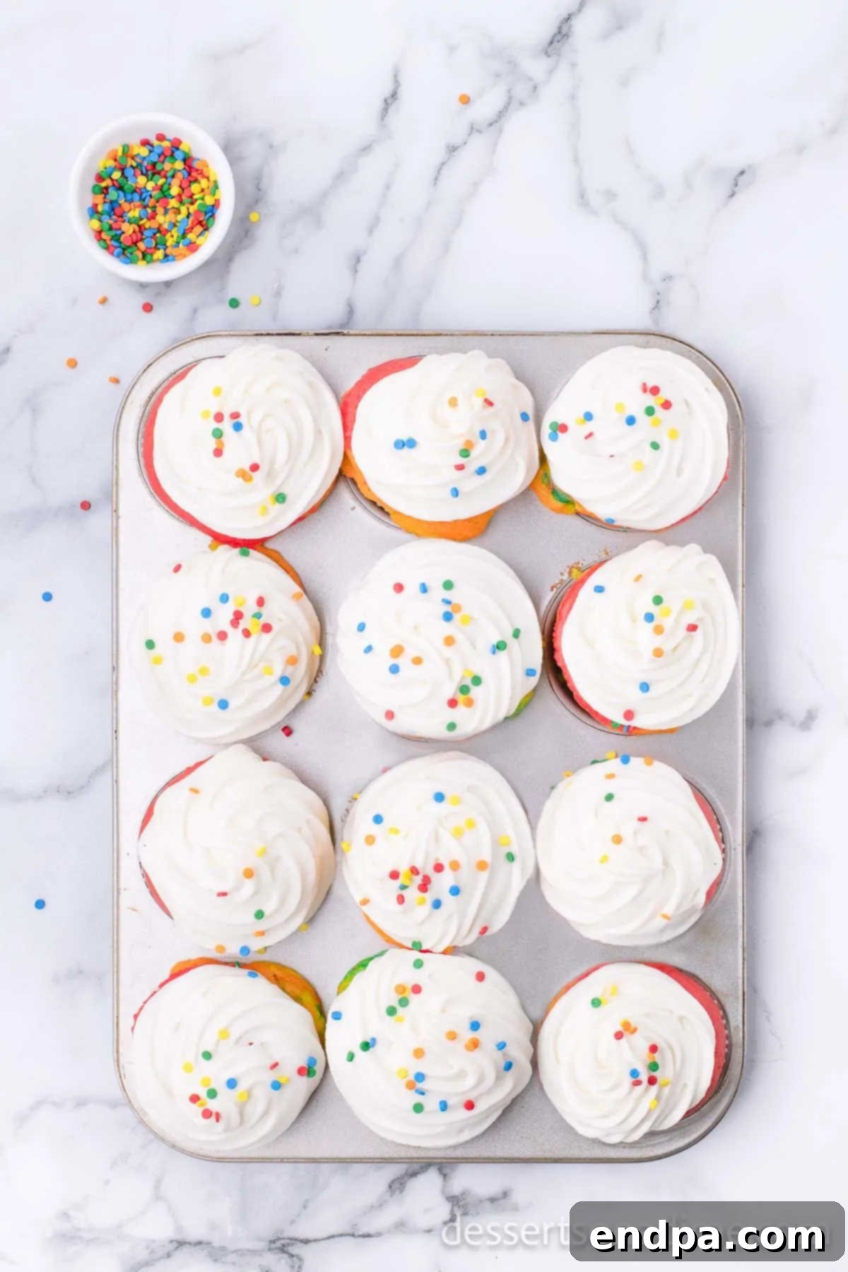 Baked rainbow cupcakes topped with fluffy white frosting and colorful sprinkles.