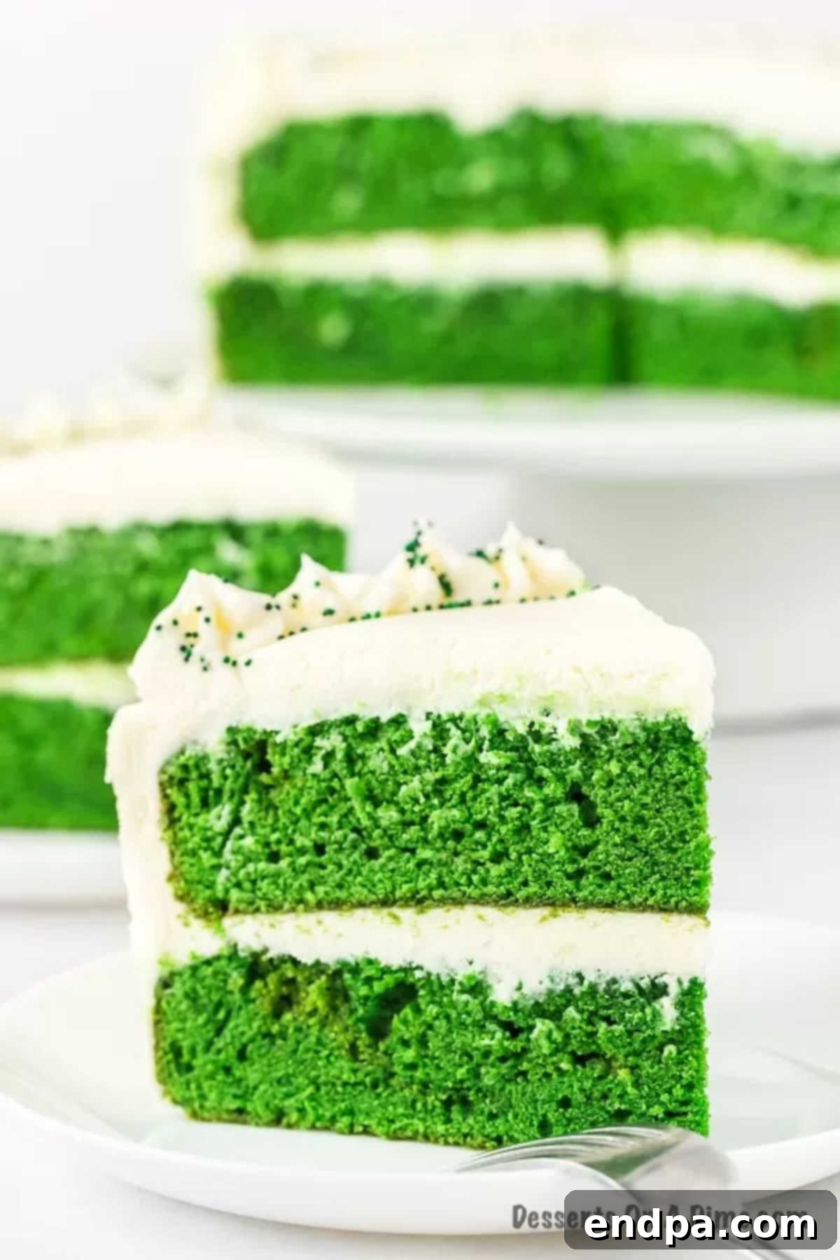 A perfectly sliced piece of Green Velvet Cake on a white plate, showcasing its vibrant green layers and thick cream cheese frosting. 