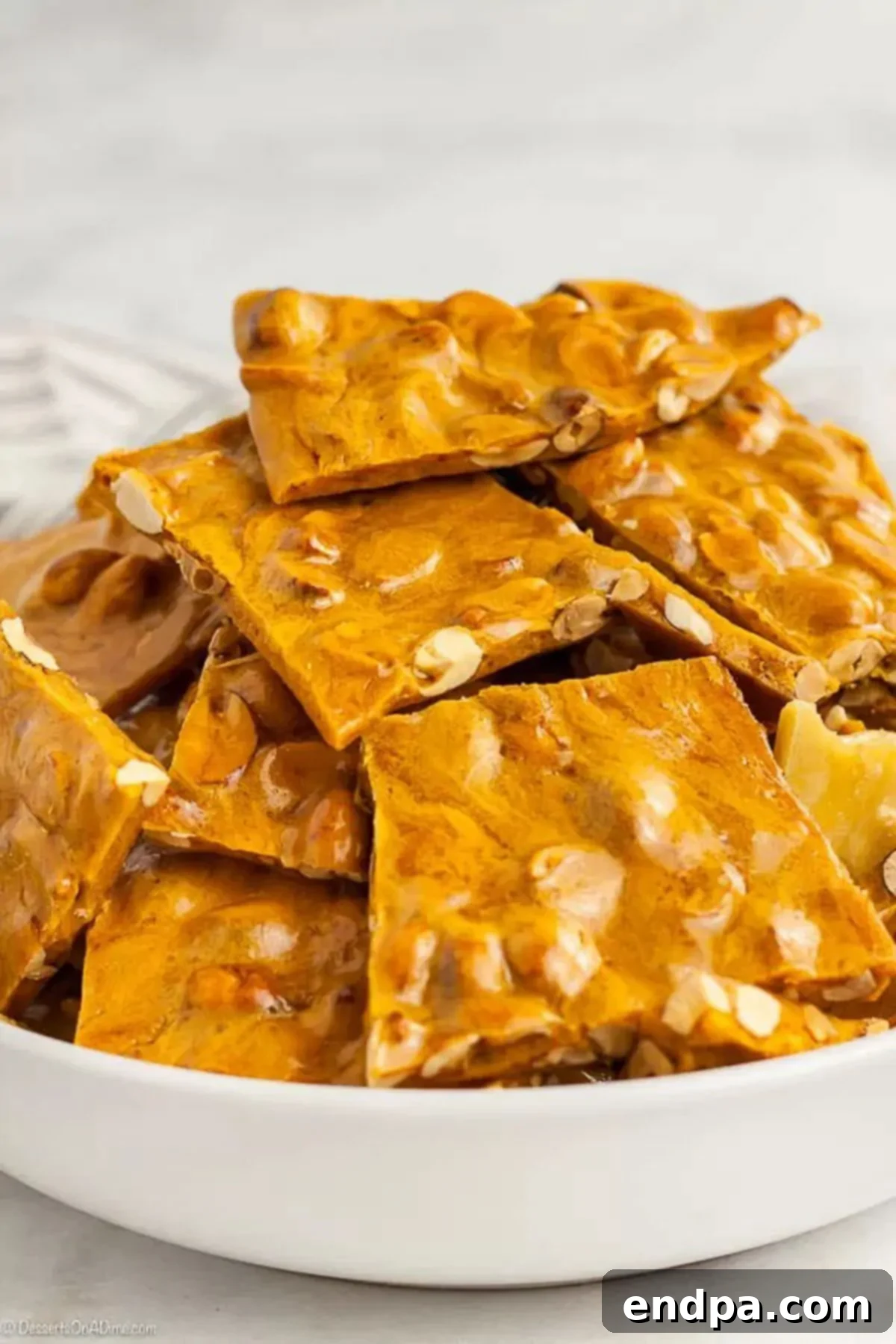 Crispy Homemade Peanut Brittle stacked neatly in a pristine white bowl, showcasing its golden hue and abundant peanuts.  