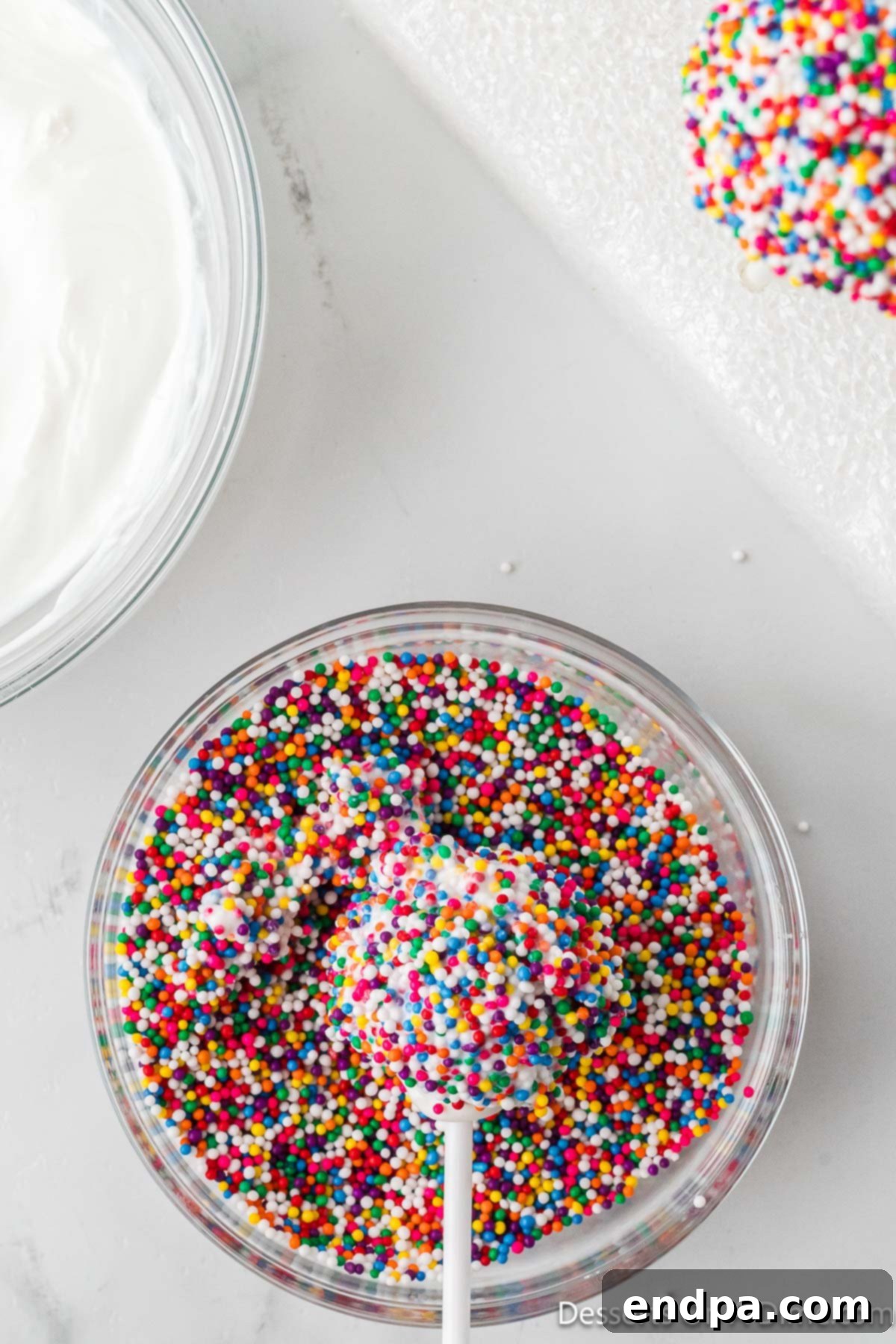 A freshly coated cake pop being rolled in a bowl of colorful rainbow sprinkles.