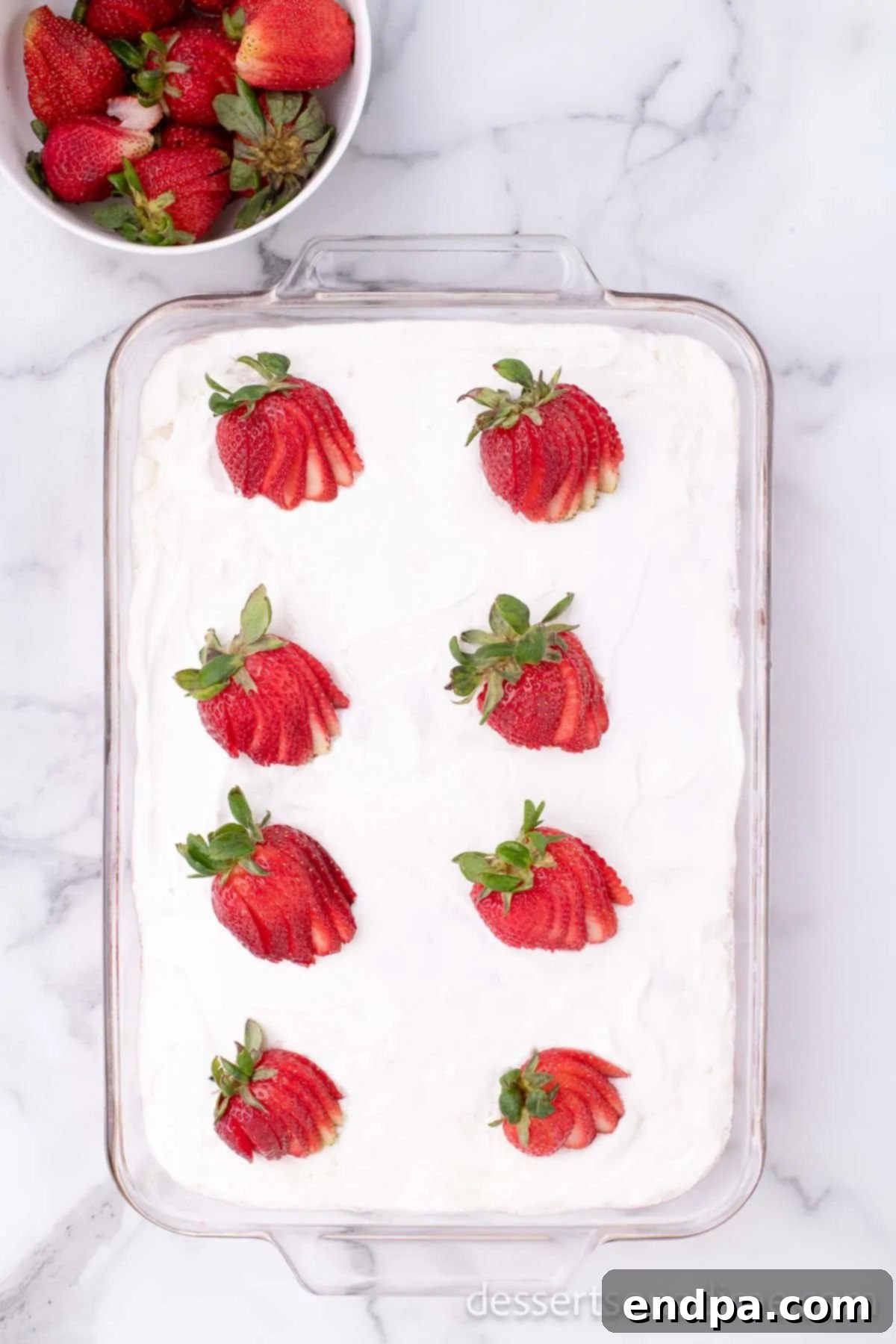 Whipped topping and fresh strawberries being added to the chilled poke cake.