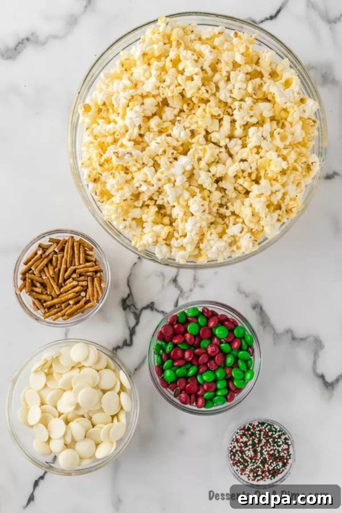 Ingredients for Christmas Crunch laid out: bags of popcorn, broken pretzels, white candy melts, red and green M&Ms, and Christmas sprinkles.