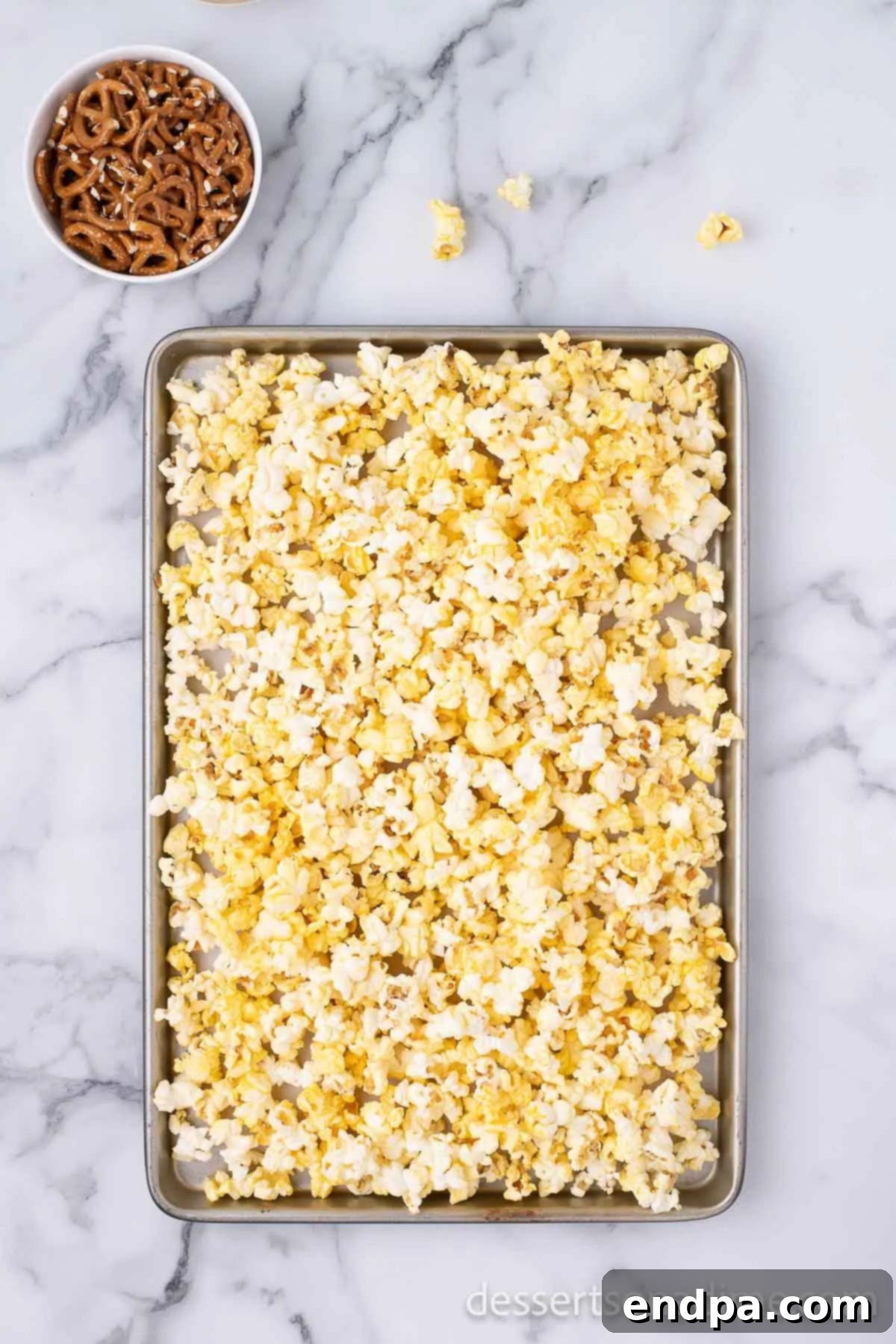 Broken pretzel pieces gently mixed into the popcorn on a baking sheet.