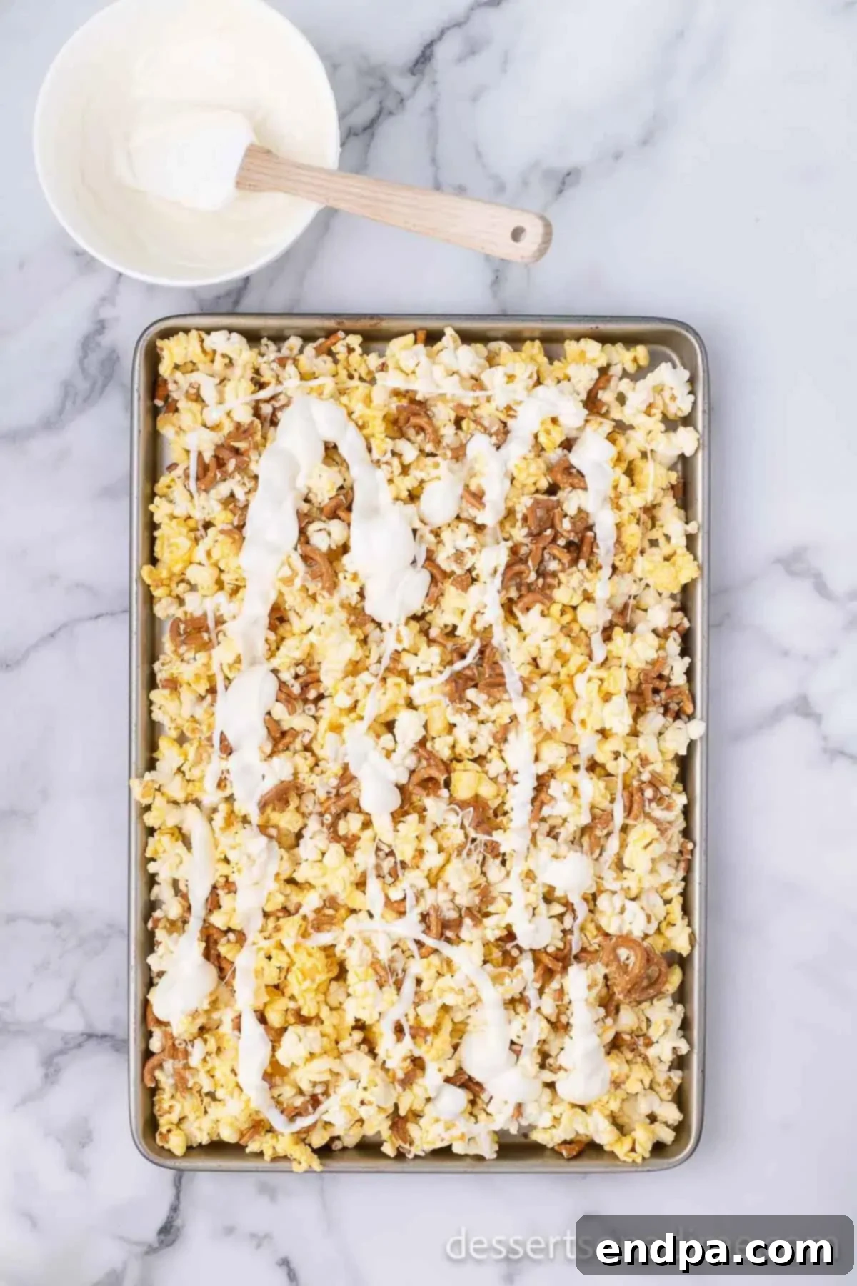 Half of the melted white chocolate drizzled over the popcorn and pretzel mixture.