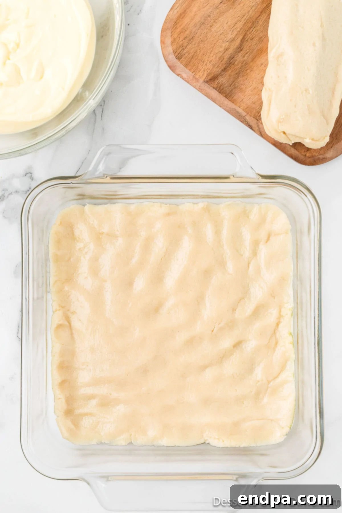 Irresistible Snickerdoodle Cheesecake Bars 6 Half of the sugar cookie dough is evenly pressed into the bottom of a baking dish, forming the golden crust for the cheesecake bars.