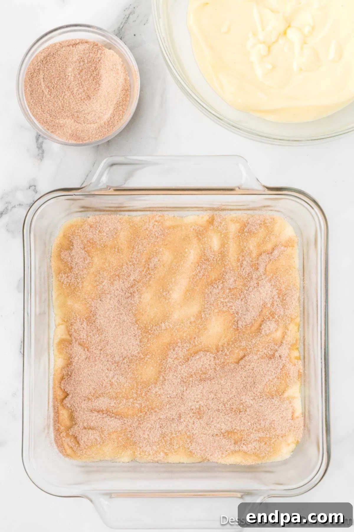 Irresistible Snickerdoodle Cheesecake Bars 8 Half of the cinnamon sugar mixture is generously sprinkled over the pressed cookie dough crust in the baking pan, preparing it for the cheesecake layer.