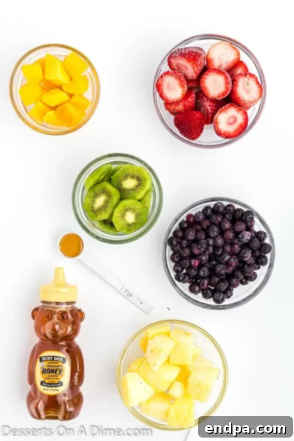 Frozen Rainbow Delights 3 A flat lay photo showing the ingredients for Rainbow Popsicles: fresh or frozen strawberries, mango chunks, pineapple chunks, peeled and sliced kiwi, blueberries, and a jar of honey.