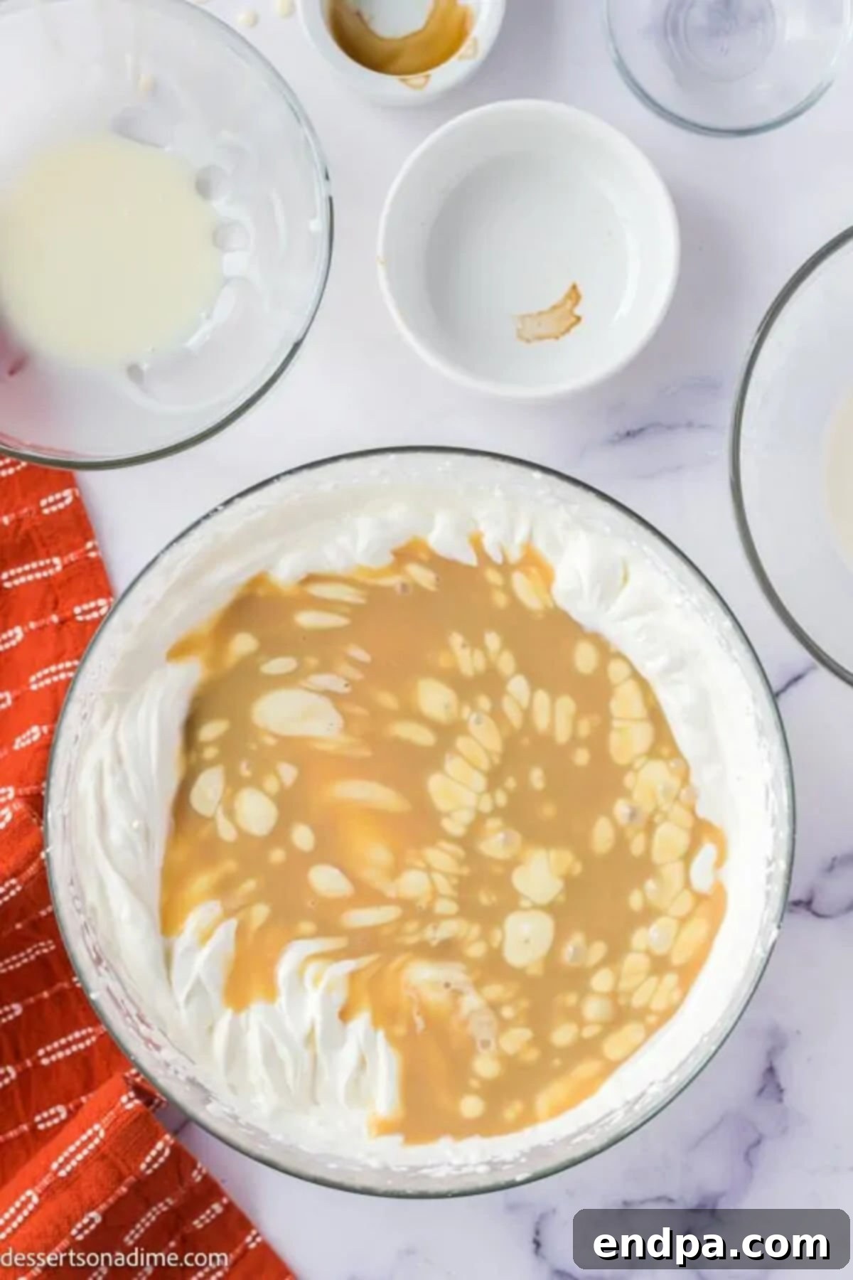 Mixing bowl containing whipped heavy cream and sweetened condensed milk, with a drizzle of butterscotch sauce being added, illustrating the combining process.