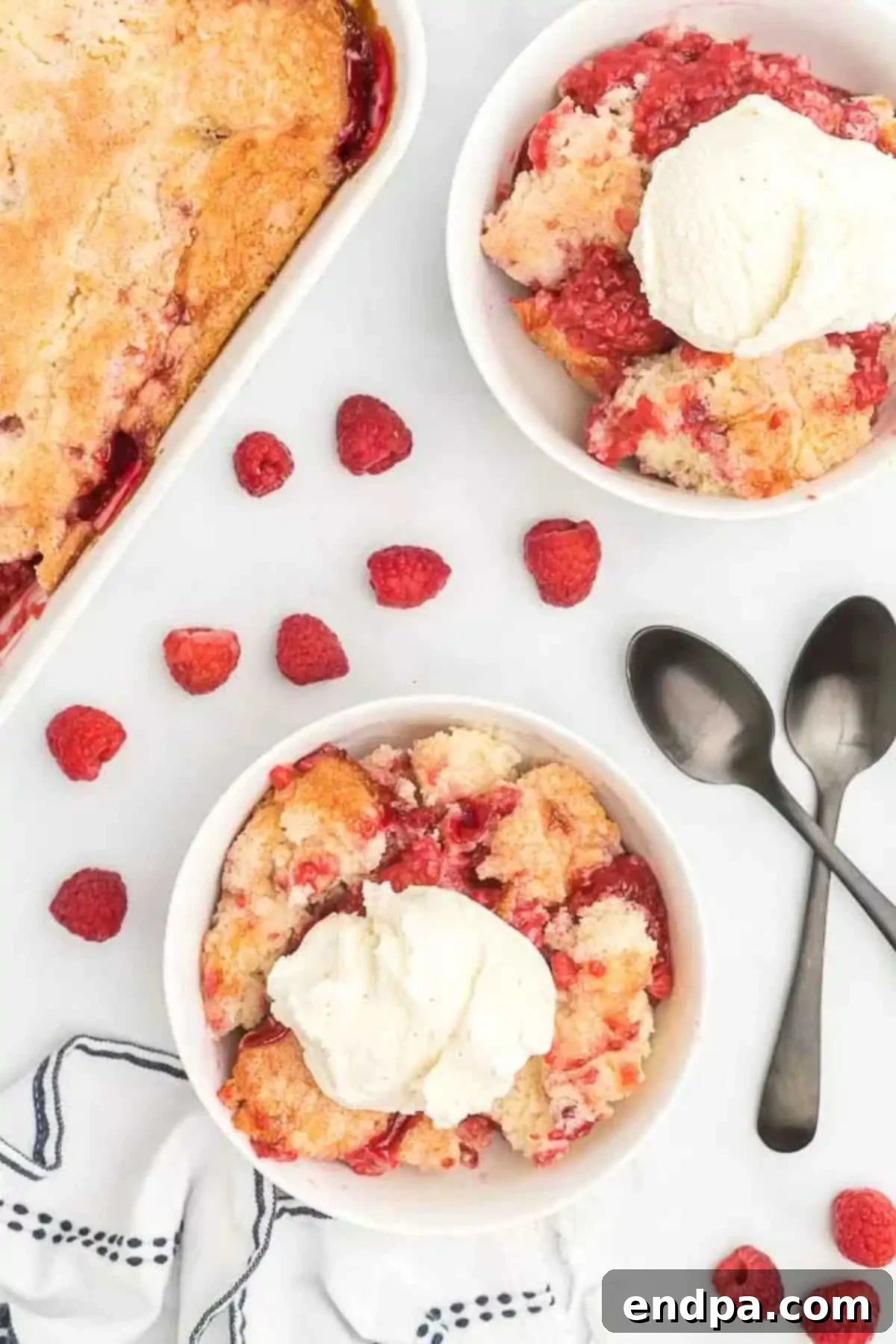 A bowl of raspberry cobbler with some fresh raspberries on the side, accompanied by two spoons.