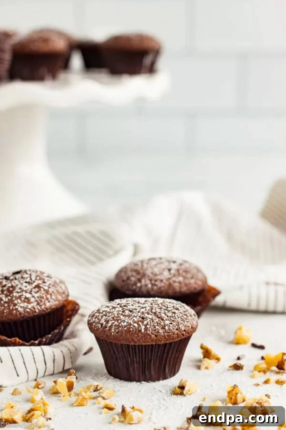 These delightful bite-sized treats are an absolute joy to eat, and this Mini Brownie Cupcakes recipe is incredibly simple to create. Each miniature brownie offers a truly decadent experience. Three Mini Brownie Cupcakes, elegantly dusted with powdered sugar, arranged on a white surface next to a scattering of delicate chocolate drizzles and sprinkles, ready to be enjoyed.