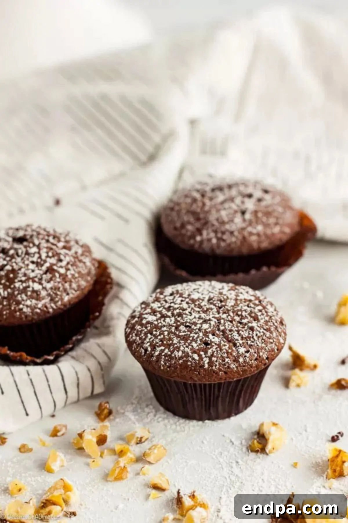 These delightful bite-sized treats are incredibly fun to eat, and this Mini Brownie Cupcakes recipe is super easy to make. Each bite is so decadent, making for an amazing mini brownie. Three Mini Brownie Cupcakes generously dusted with powdered sugar, arranged on a white surface with scattered nuts and chocolate drizzles, highlighting their bite-sized perfection.