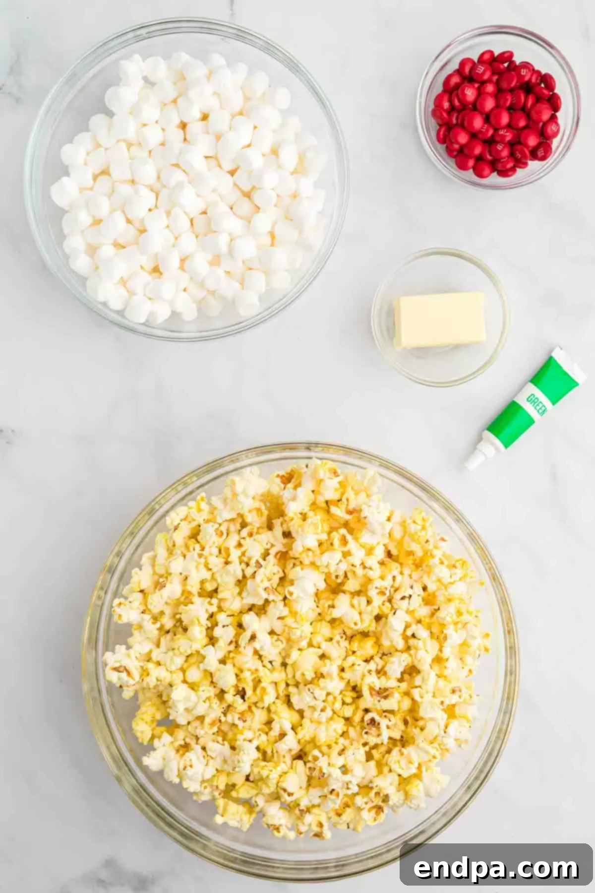 Festive Grinch Popcorn Mix 3 Grinch Popcorn ingredients including cooked popcorn, butter, mini marshmallows, green gel food coloring, and red M&Ms, laid out on a table.