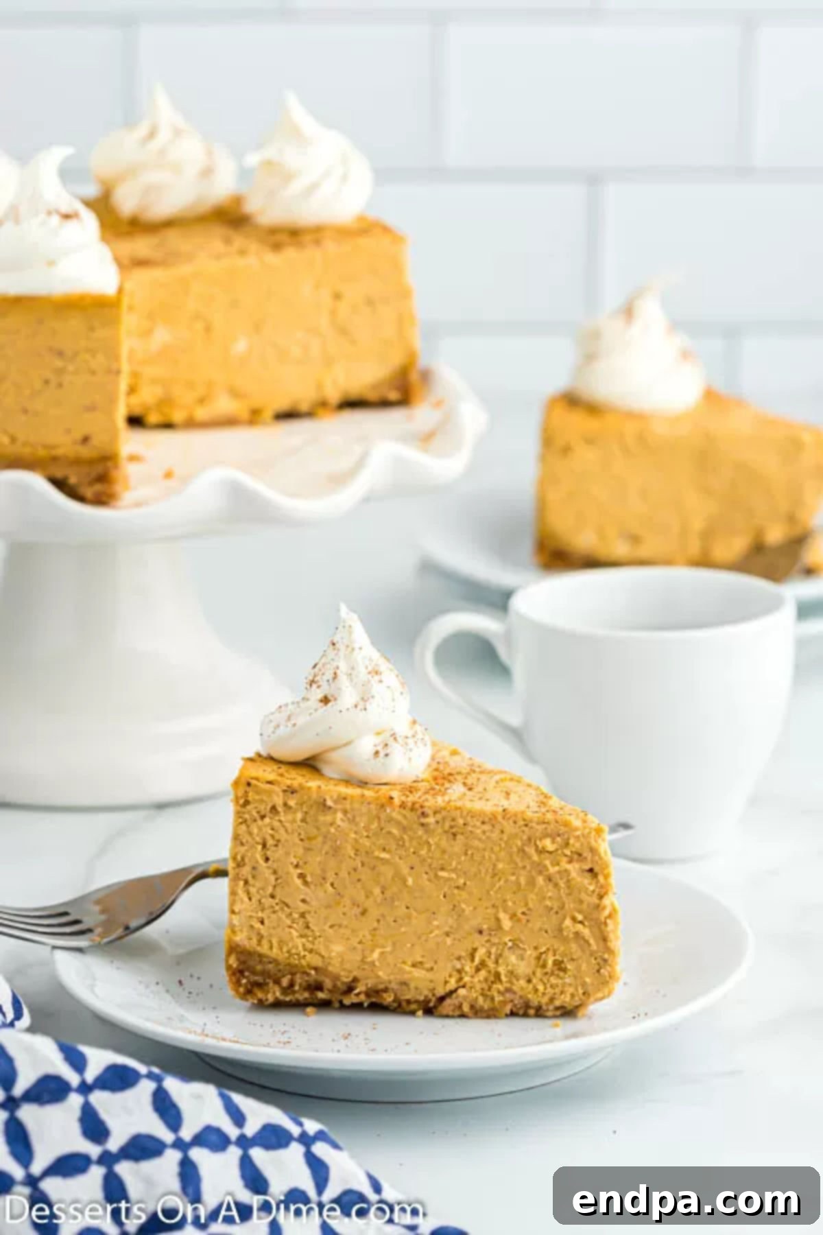 A slice of Instant Pot Pumpkin Cheesecake on a plate, with the rest of the cheesecake on a stand in the background, topped with whipped cream.