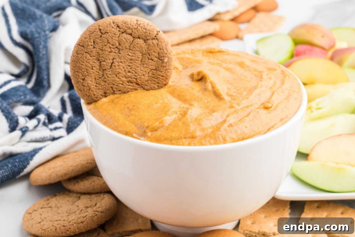 Creamy pumpkin dip in a elegant white bowl, surrounded by various cookies and fresh fruit, ready to be served for a fall gathering.