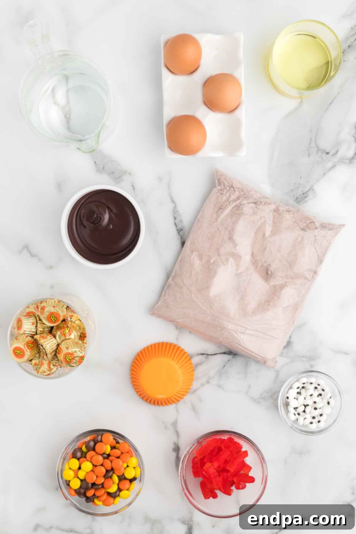 A flat lay of all the simple ingredients required to make delicious Turkey Cupcakes, including chocolate cake mix, orange cupcake liners, chocolate frosting, Reese's Peanut Butter Cups, candy eyes, and Reese's Pieces.
