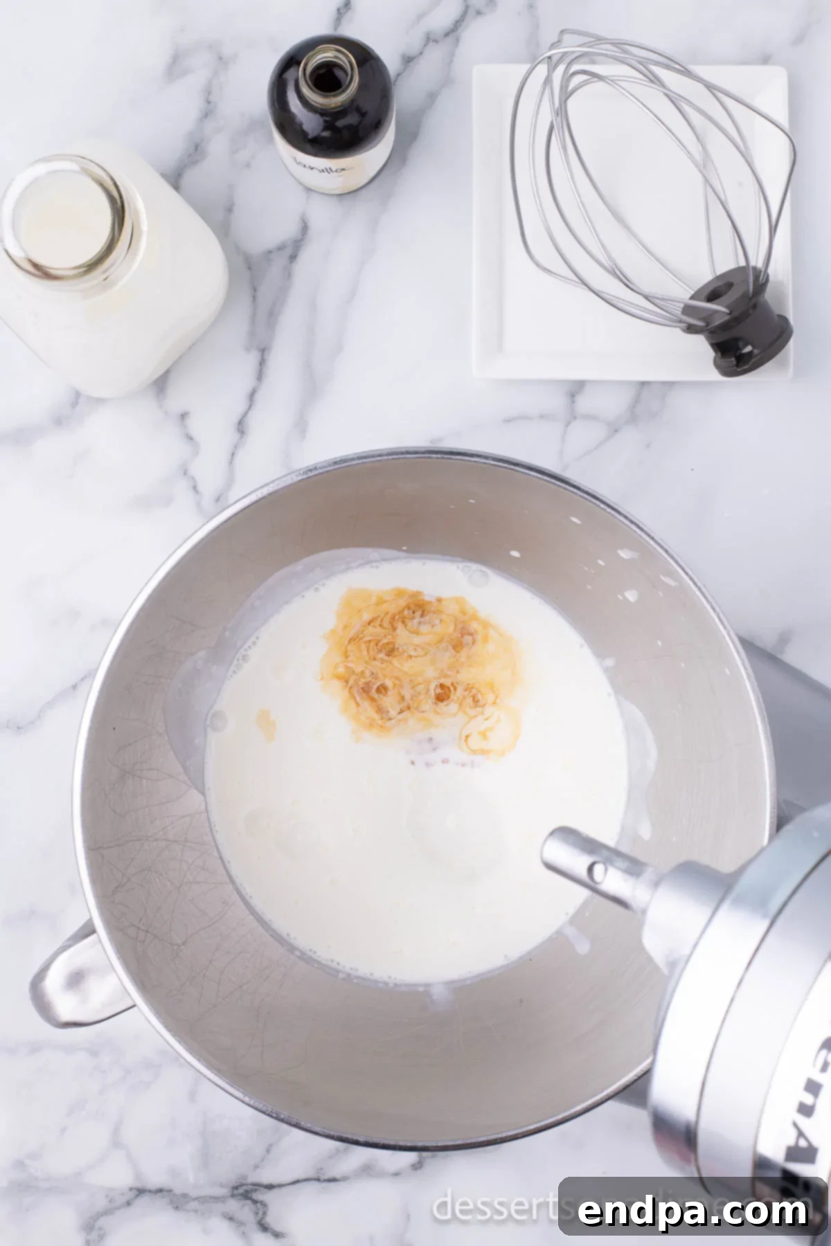 Mixing bowl with heavy cream and vanilla extract being combined by an electric mixer.