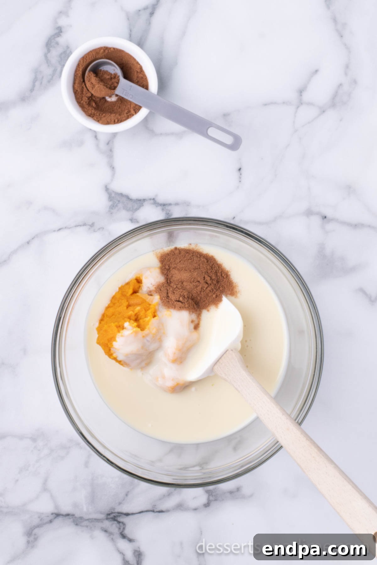 Pumpkin pie spice being sprinkled into the mixing bowl containing pumpkin puree and sweetened condensed milk.