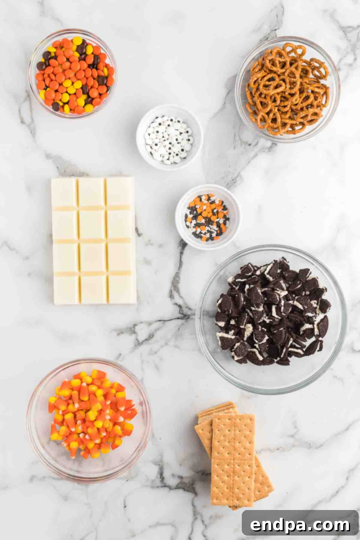 A flat lay of Halloween Candy Bark ingredients: graham crackers, white almond bark, chopped Oreos, Reese's Pieces, Halloween sprinkles, candy eyes, mini pretzel twists, and candy corn.