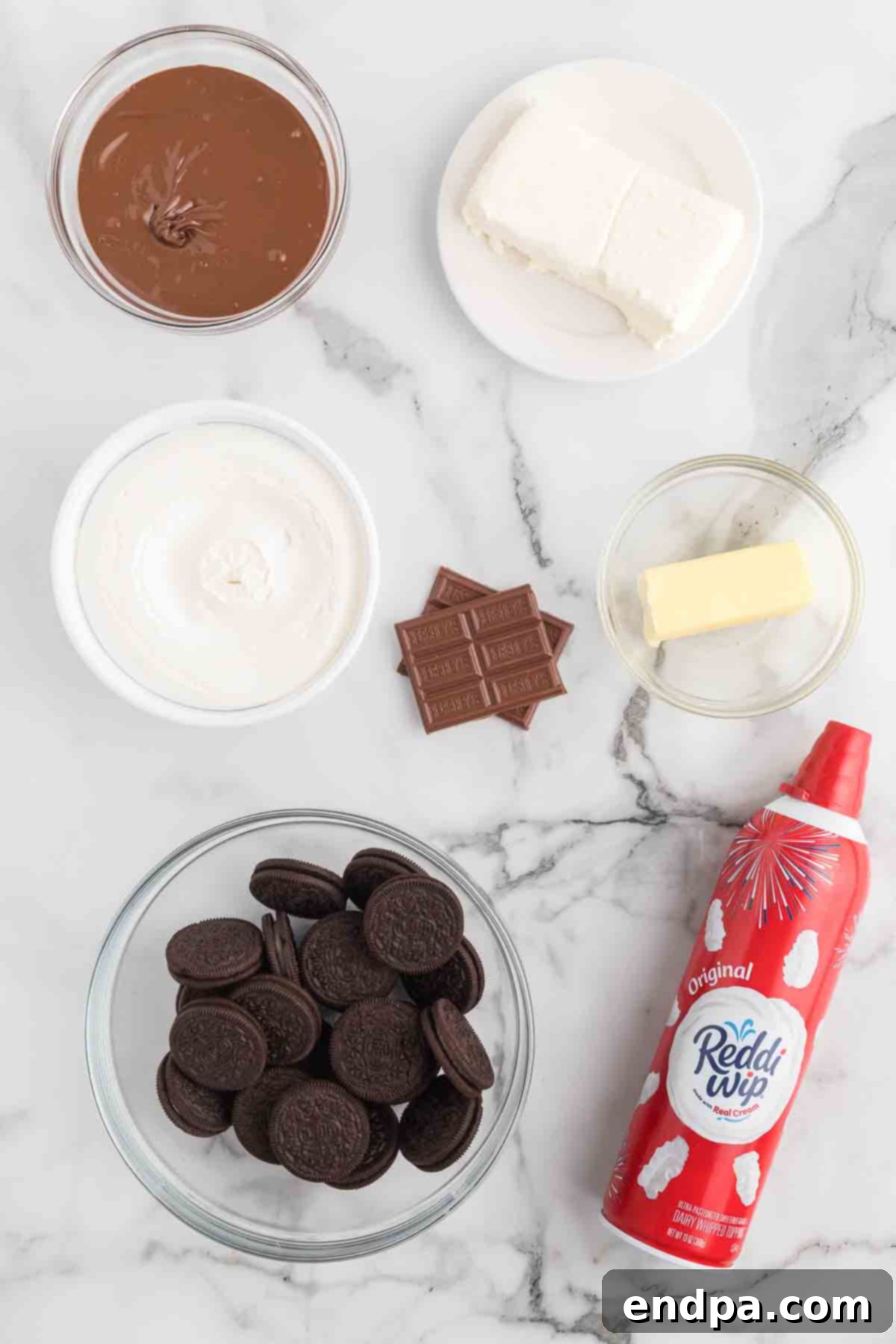 A collection of ingredients laid out for making Nutella Pie, including Oreo cookies, butter, cream cheese, Nutella, whipped topping, and a milk chocolate bar.