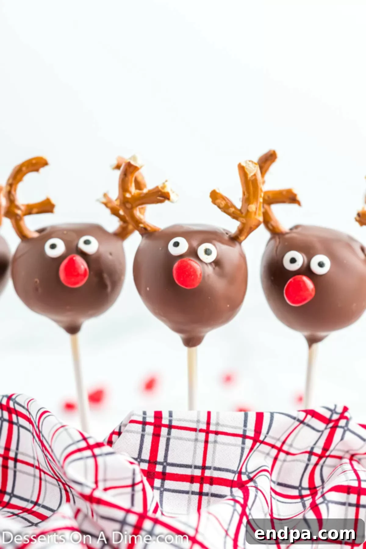 A festive Reindeer Cake Pop, standing elegantly and showcasing its charming pretzel antlers, candy eyes, and a bright red nose against a blurred background.