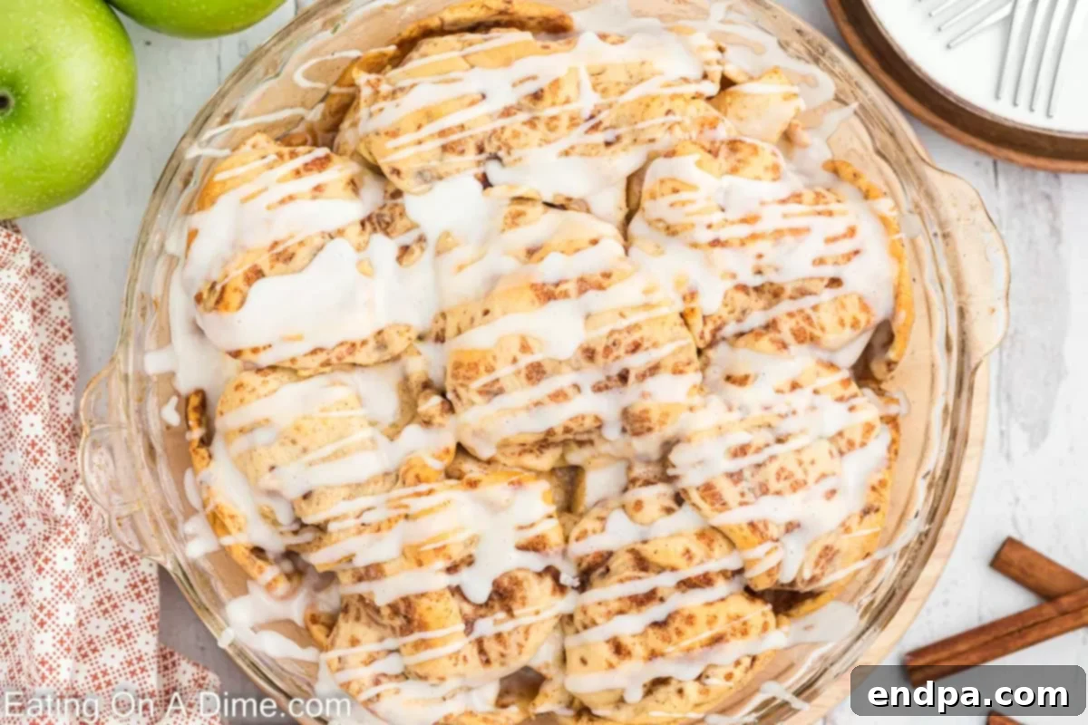 A beautifully baked Cinnamon Roll Apple Pie, drizzled with sweet icing, ready to be served from a ceramic pie dish. The golden-brown crust made from cinnamon rolls peeks through.