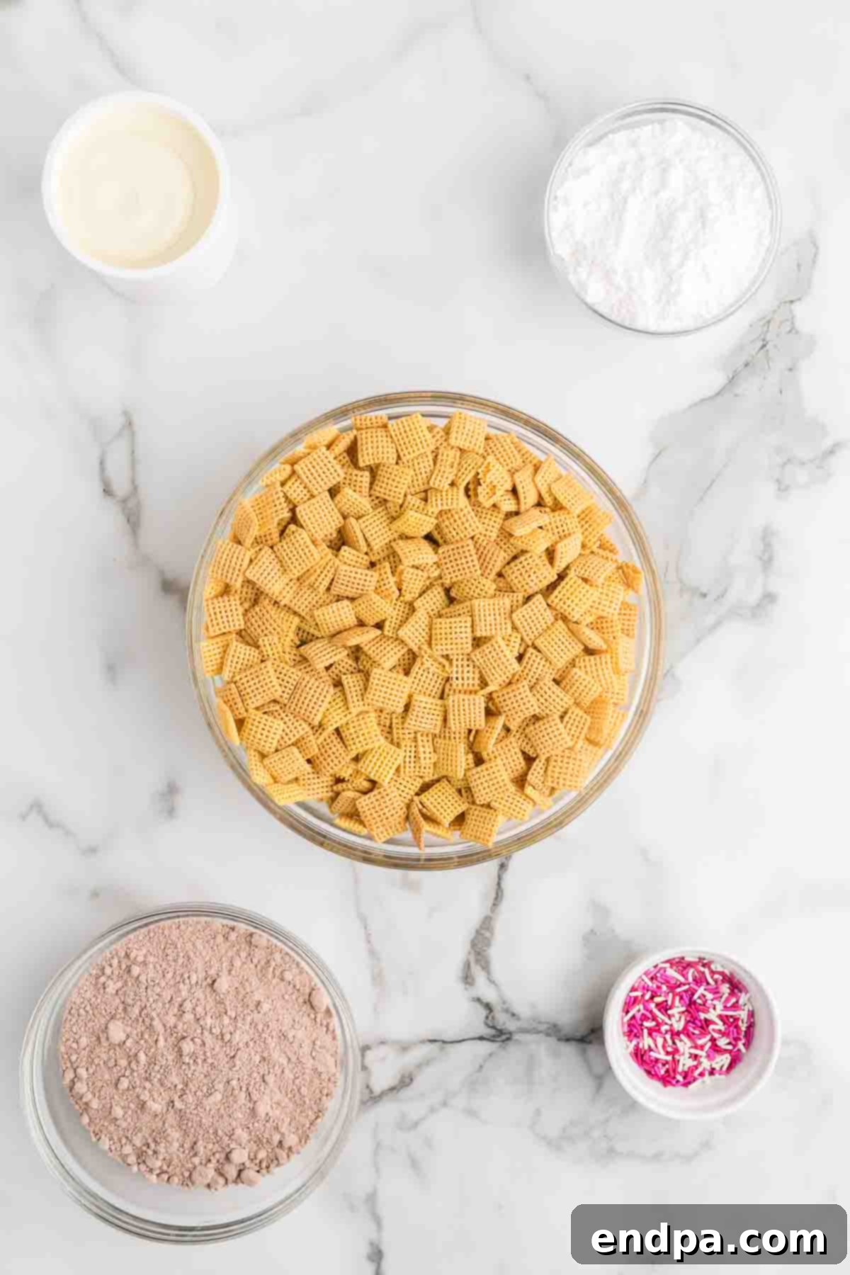 Ingredients for red velvet puppy chow spread out on a table: Chex cereal, cream cheese frosting, red velvet cake mix, powdered sugar, and an assortment of red, white, and pink sprinkles.
