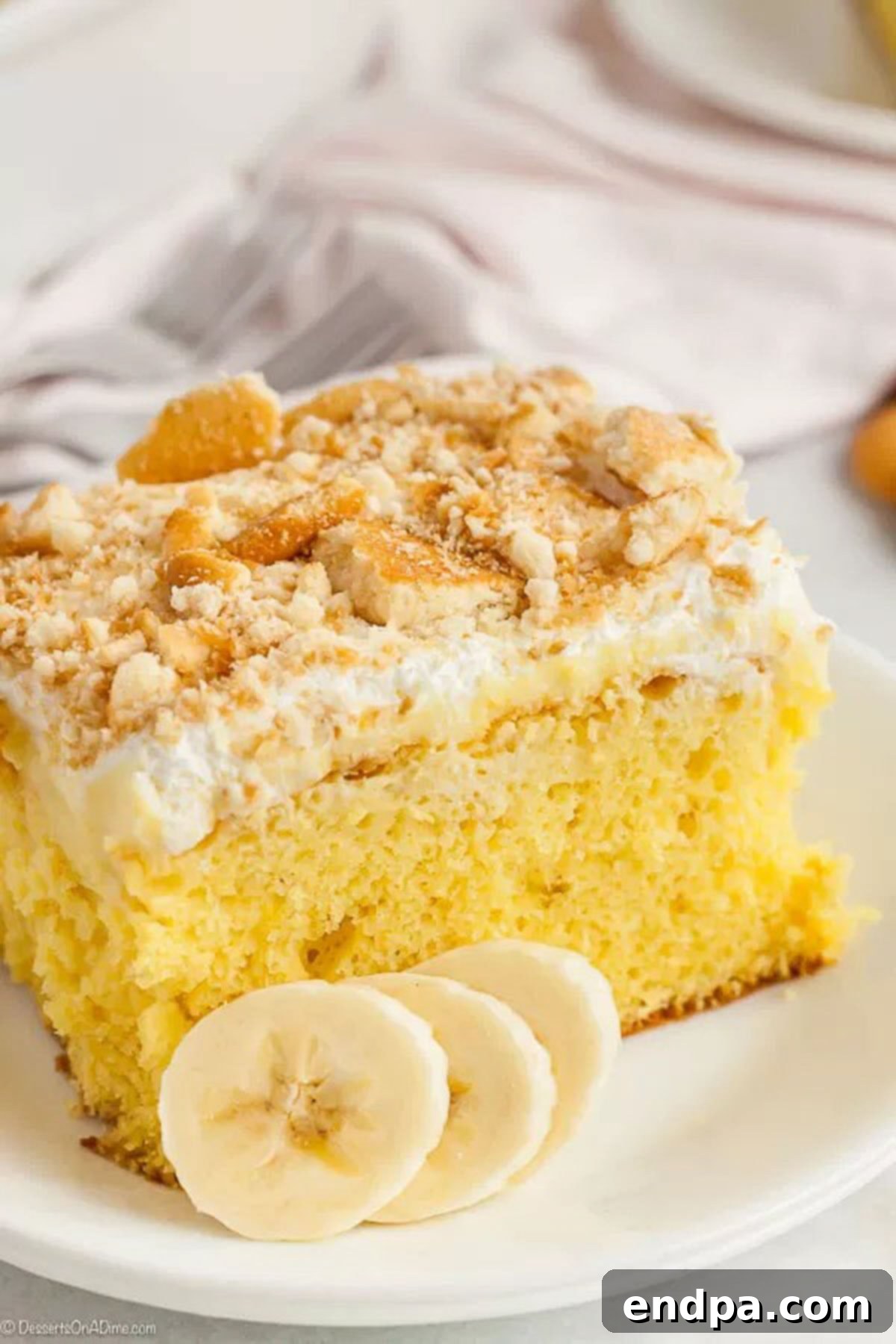 Banana Dream Pudding Cake 2 A slice of this banana pudding poke cake on a white plate topped with crushed vanilla wafers and sliced bananas next to the cake on the plate