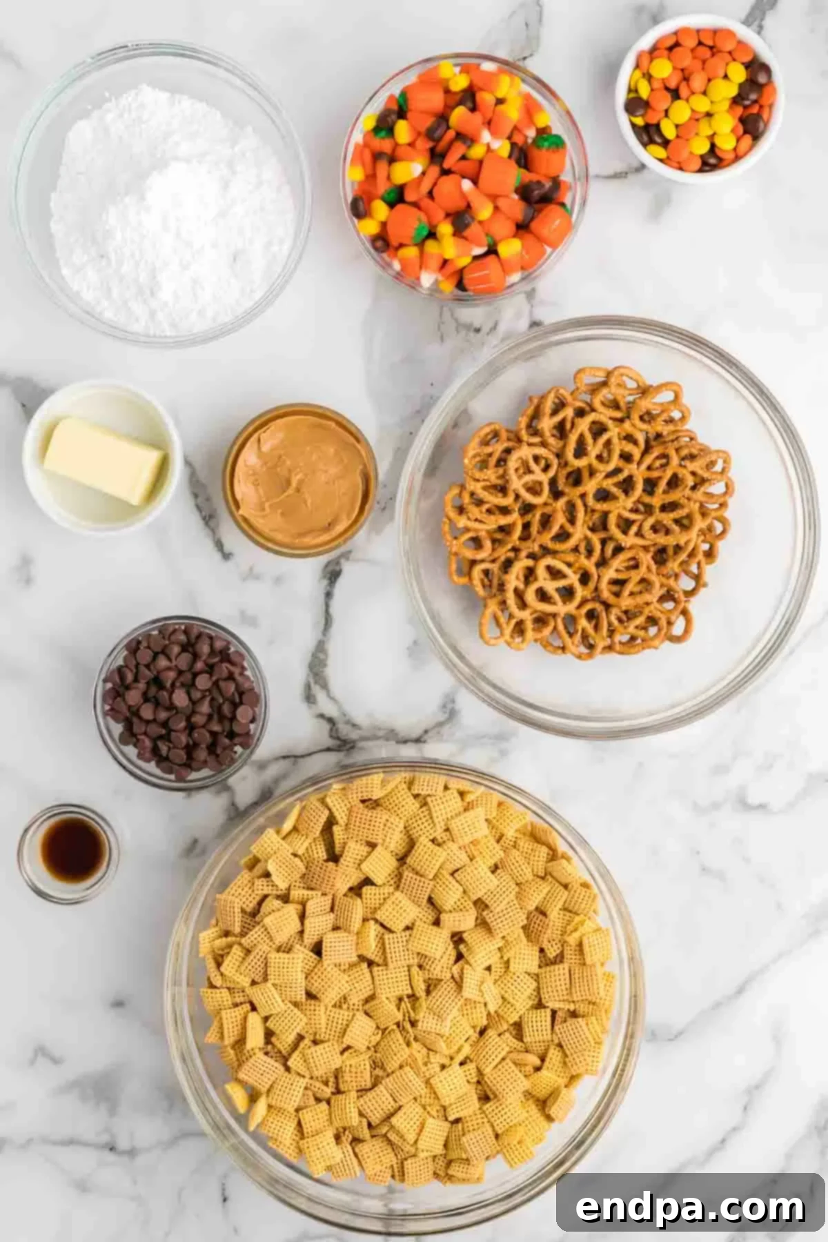 A selection of ingredients laid out on a table, including chocolate chips, peanut butter, butter, vanilla, Chex cereal, powdered sugar, autumn mix candy, pretzels, and Reese's Pieces.