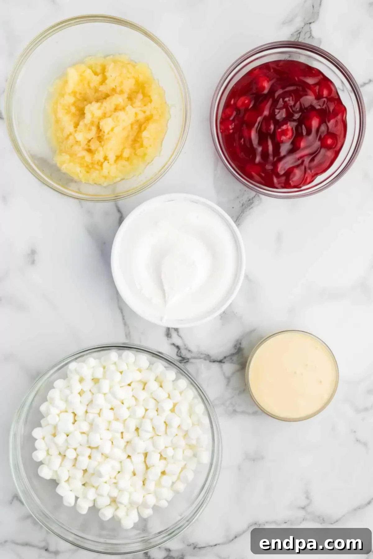 A spread of ingredients laid out, featuring cans of cherry pie filling, sweetened condensed milk, crushed pineapple, and bags of mini marshmallows and whipped topping. 