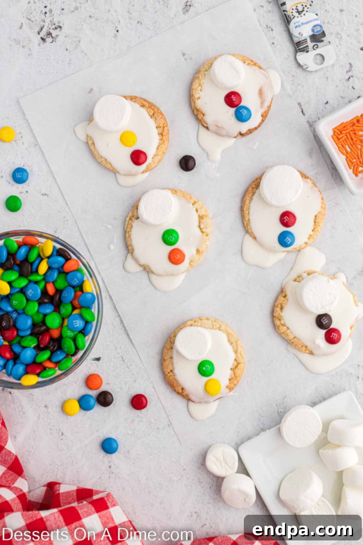 Whimsical Melted Snowman Treats 6 M&M's placed on each cookie. Partially frosted cookies are shown with colorful M&Ms being carefully positioned on the lower half of the cookie as snowman buttons, with space reserved for the marshmallow head.