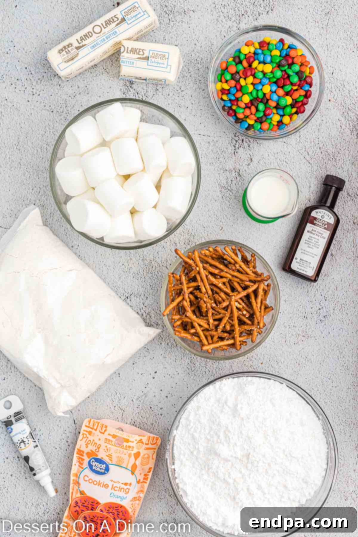 A flat lay of ingredients for snowman cupcakes including a cake mix box, colorful M&M's, large marshmallows, pretzel sticks, powdered sugar, and a stick of butter.
