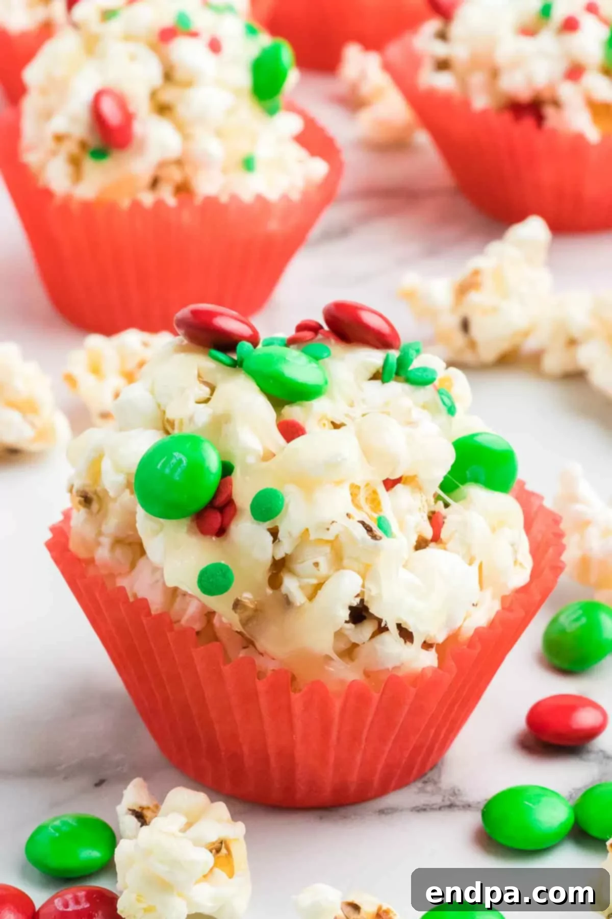 Holiday Popcorn Ball Delight 8 A close-up photograph showcasing three vibrant Christmas popcorn balls, each nestled charmingly in a red paper muffin liner, highlighting their festive colors and sprinkles.