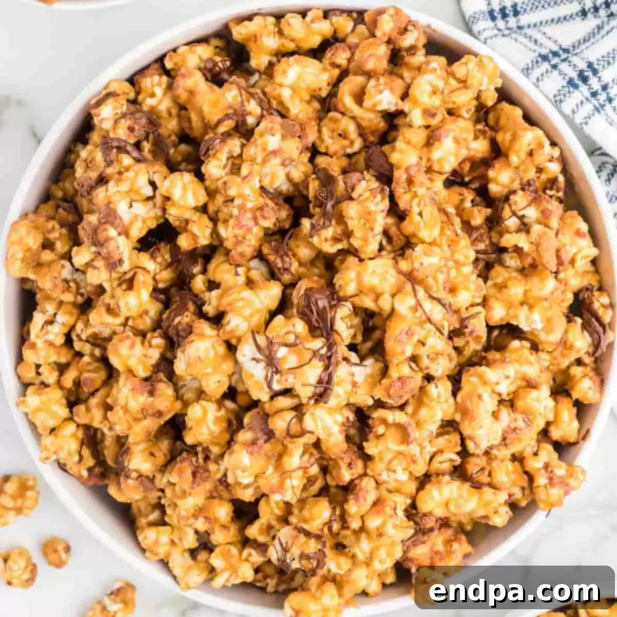 A close-up image of Snickers popcorn in a white bowl, showing individual kernels coated in caramel, studded with Snickers pieces, and drizzled with chocolate.