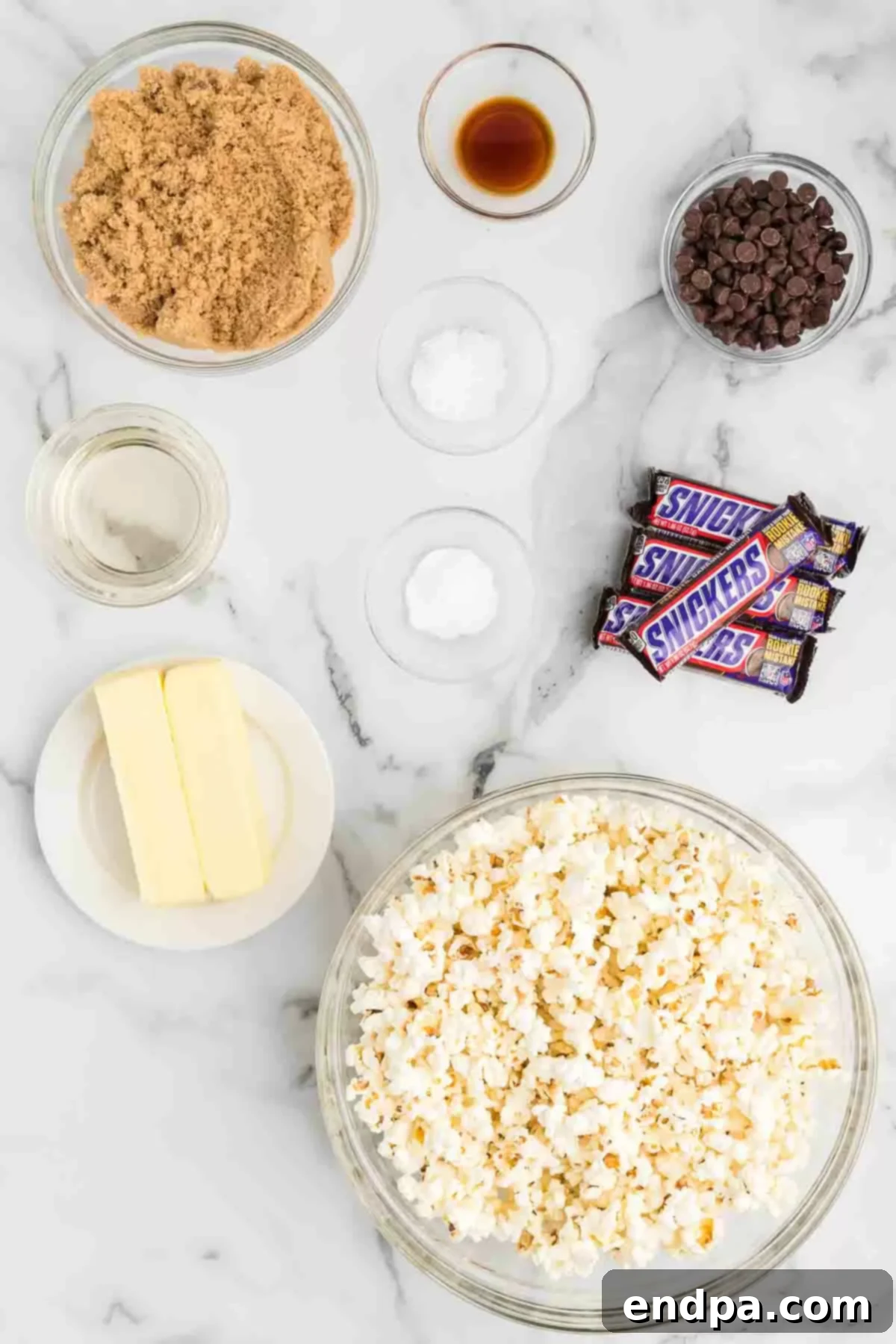 A visual display of all ingredients needed for Snickers popcorn, including popped popcorn, butter, brown sugar, baking soda, vanilla extract, Snickers bars, and semi-sweet chocolate chips.