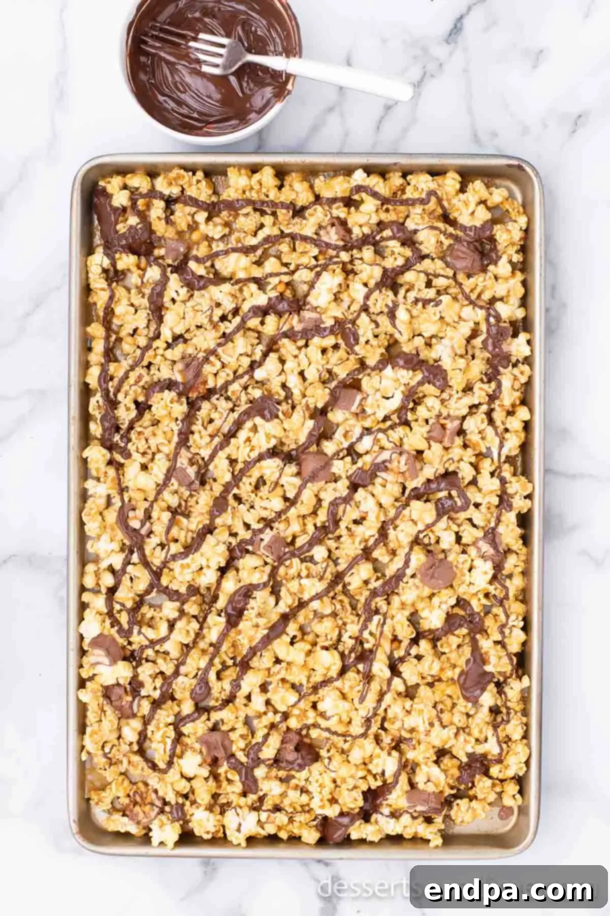 Melted semi-sweet chocolate being drizzled in thin streams over the cooled Snickers caramel popcorn.