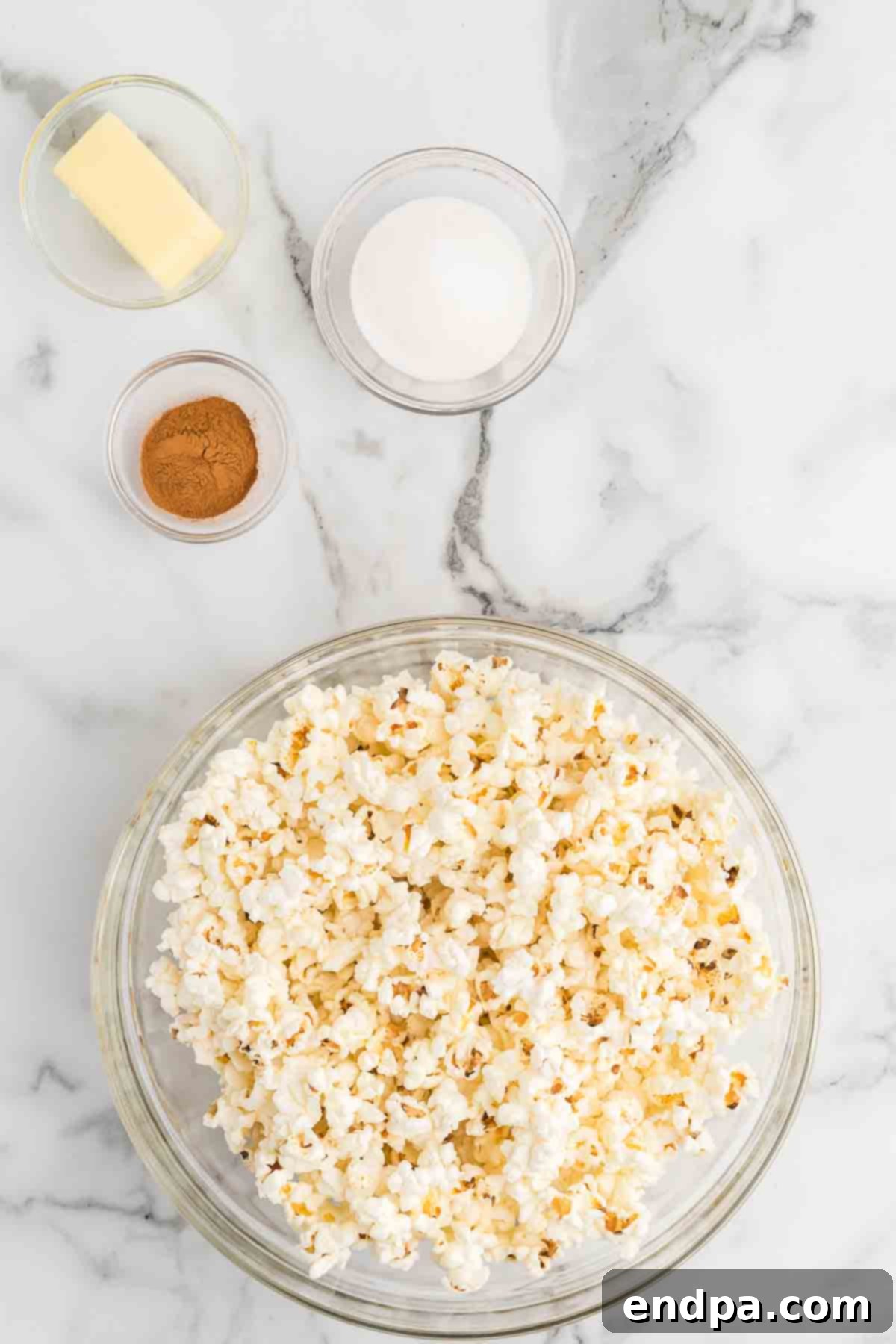 A spread of ingredients for Cinnamon Toast Crunch Popcorn: a bowl of freshly popped popcorn, a stick of butter, a bowl of granulated sugar, and a small jar of ground cinnamon.