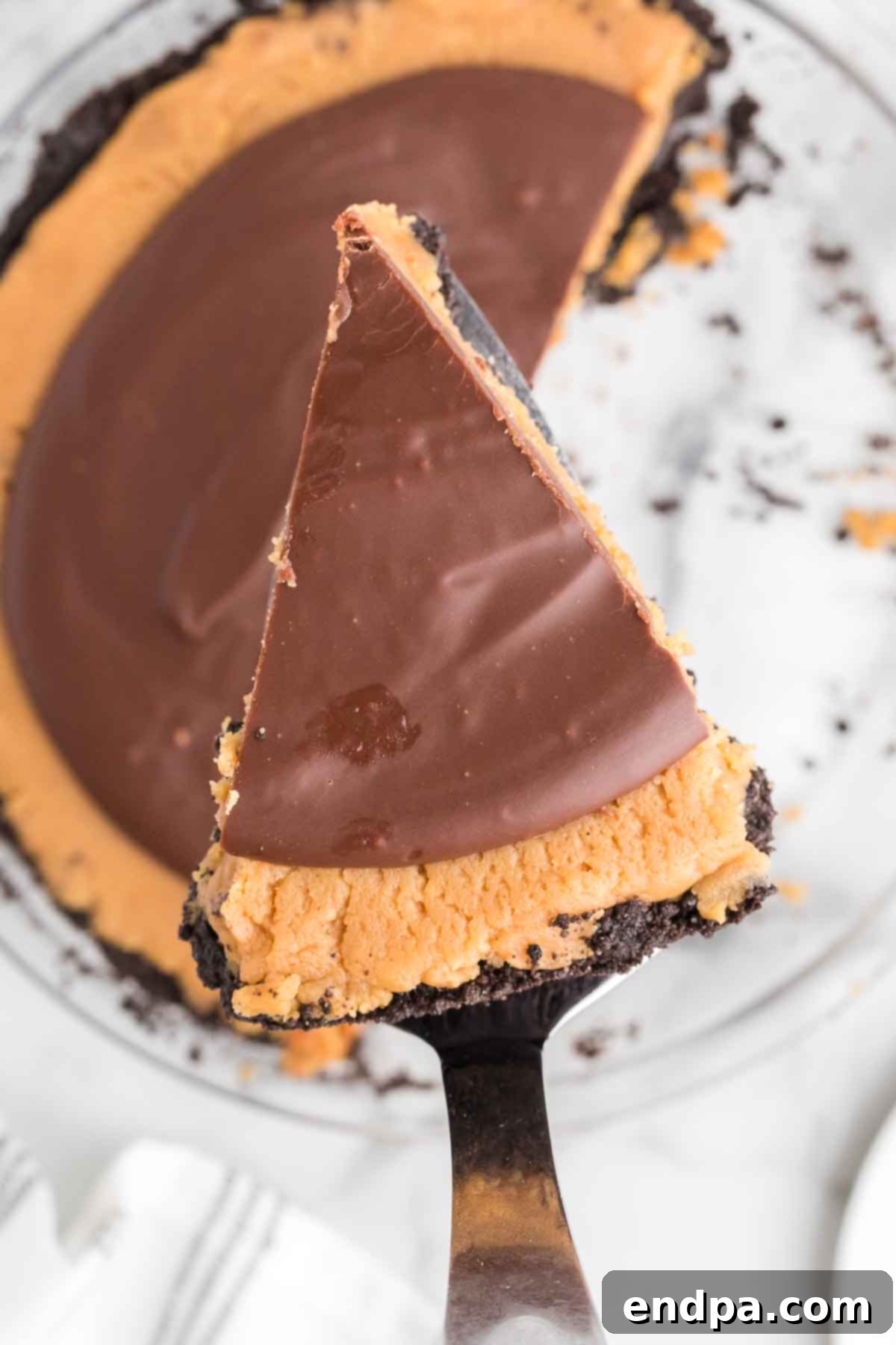 A beautifully presented slice of homemade Buckeye pie on a white plate, showcasing its distinct layers of dark Oreo crust, light peanut butter filling, and glossy chocolate ganache topping. The pie is partially sliced in the background, revealing more of its tempting interior.