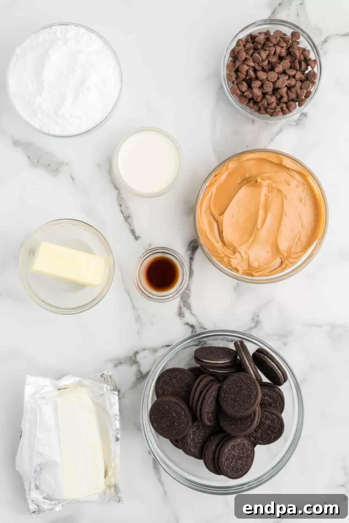 A collection of ingredients laid out for making a Buckeye pie, including a stack of Oreo cookies, a stick of butter, a block of cream cheese, a jar of creamy peanut butter, a bag of powdered sugar, a bottle of vanilla extract, and bowls of milk chocolate chips and heavy whipping cream.