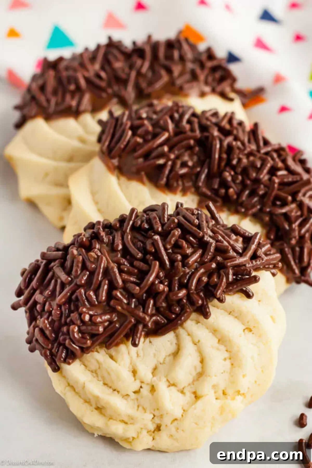 With just a few ingredients, Butter Cookies Recipe comes together quickly for a tasty and gorgeous treat. Easy butter cookies are so pretty and delicious. Two golden-brown butter cookies, beautifully dipped in chocolate frosting and adorned with chocolate sprinkles, presented on a pristine white surface.