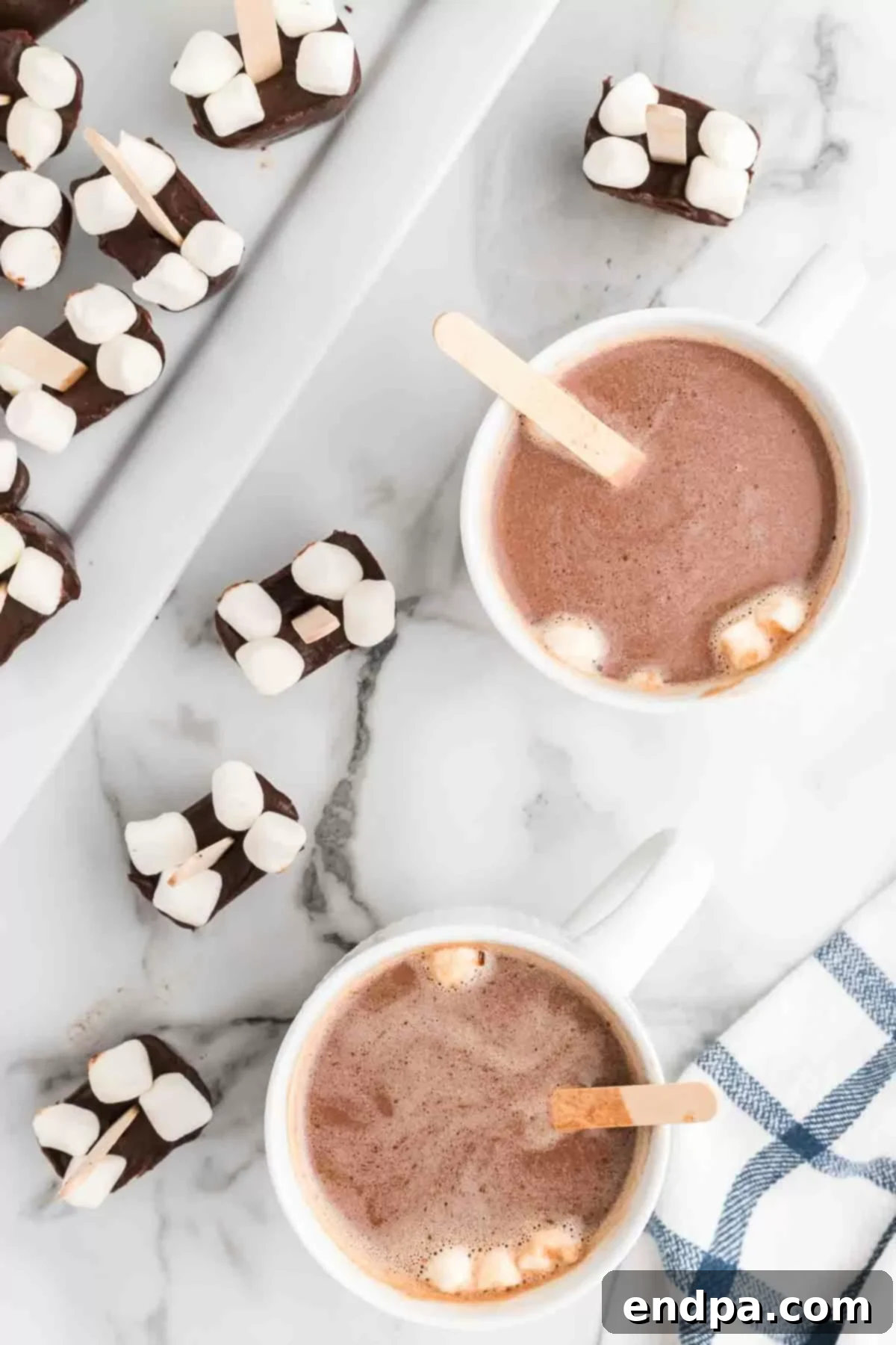 Two mugs of hot cocoa, one with a hot chocolate stick in it, topped with whipped cream and chocolate shavings.